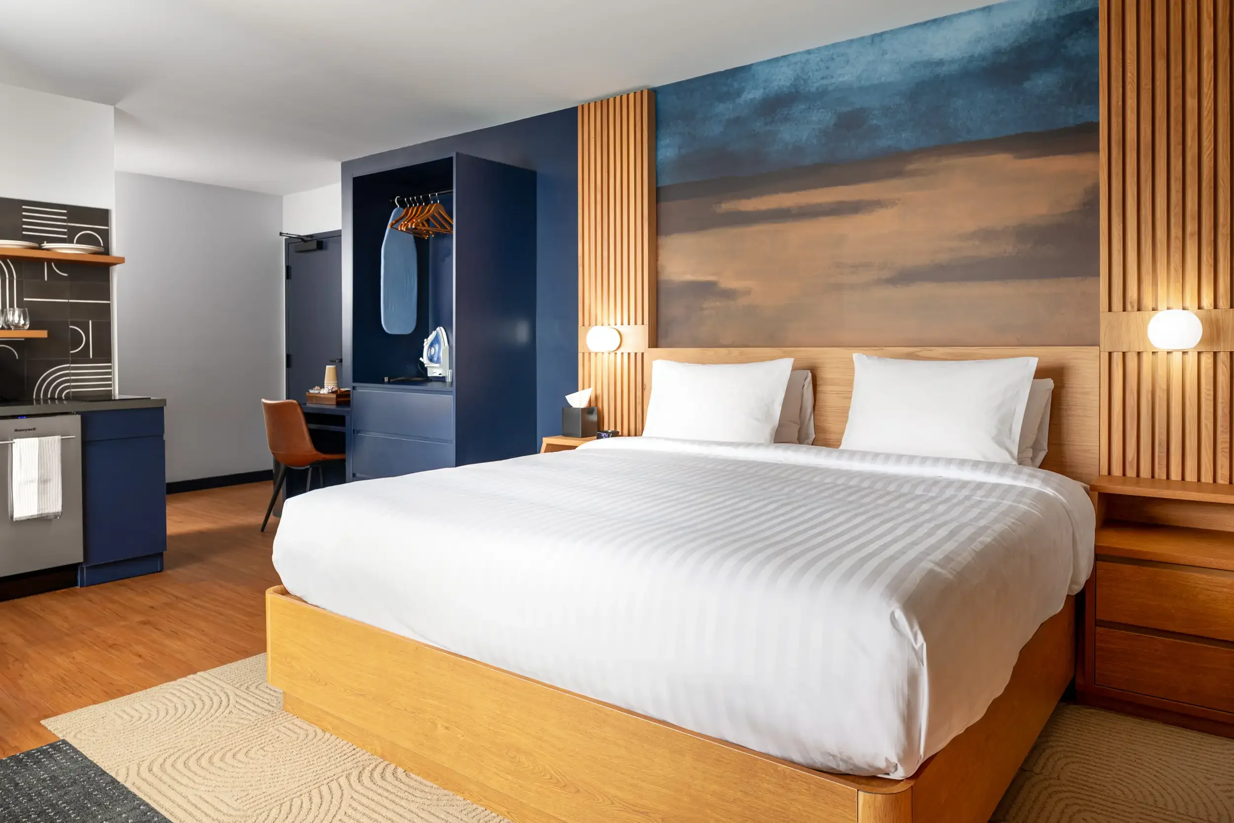 A modern hotel room with a large bed, wooden headboard and side tables, blue accent wall, and a mural of a landscape behind the bed. There is a small workspace with a desk, chair, and lamp, and a closet with an iron and hangers. The room features woo