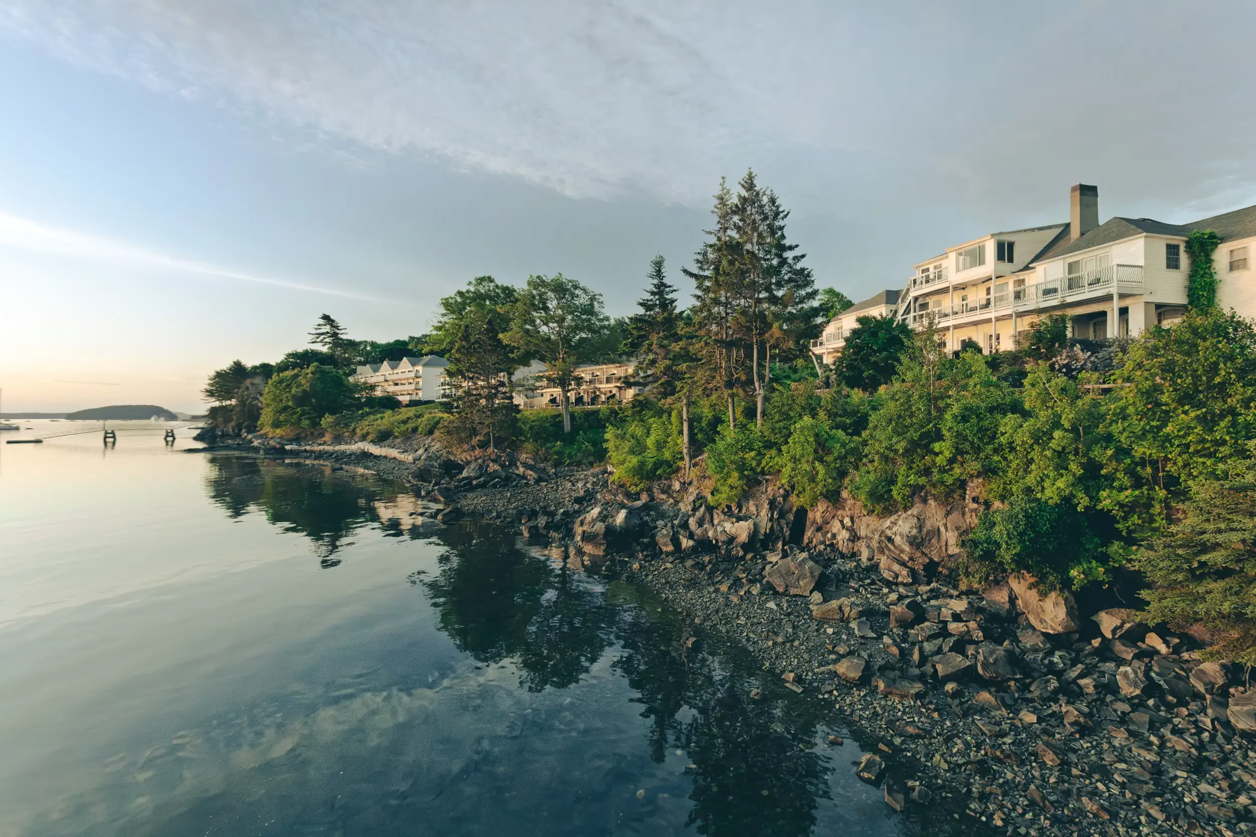 A scenic riverside view with houses on a hill surrounded by trees, calm water reflecting the sky, and a distant island with a sunset or sunrise sky.