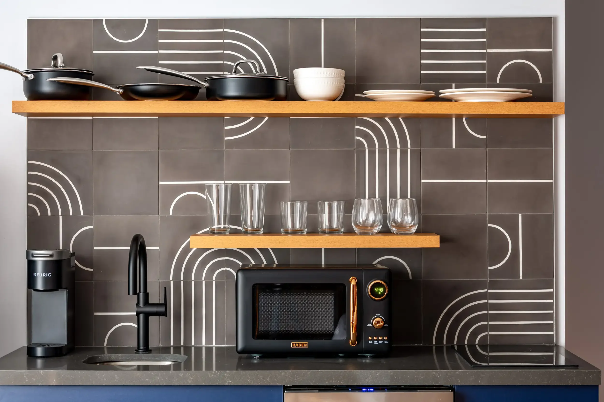 A modern kitchen with black and gray tiles featuring white geometric patterns, two wooden shelves holding white bowls, plates, and glassware, a black faucet over the sink, a black microwave oven, and a Keurig coffee machine on the counter.