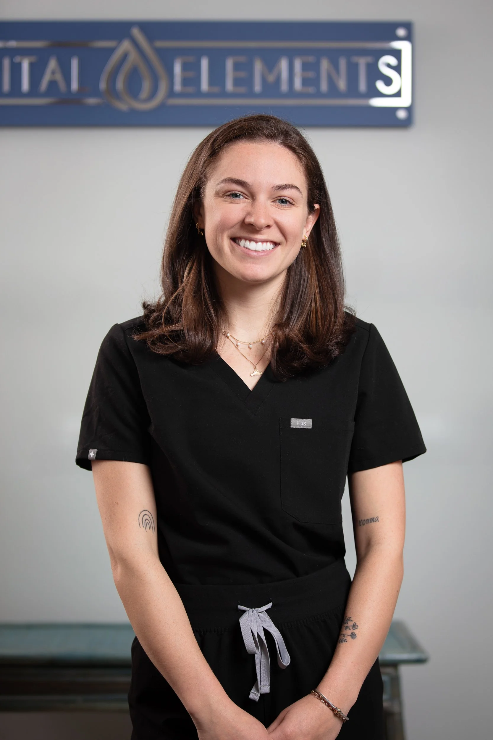 A young woman with brown hair, wearing black medical scrubs and jewelry, standing in front of a sign that reads 'Vital Elements', smiling at the camera.