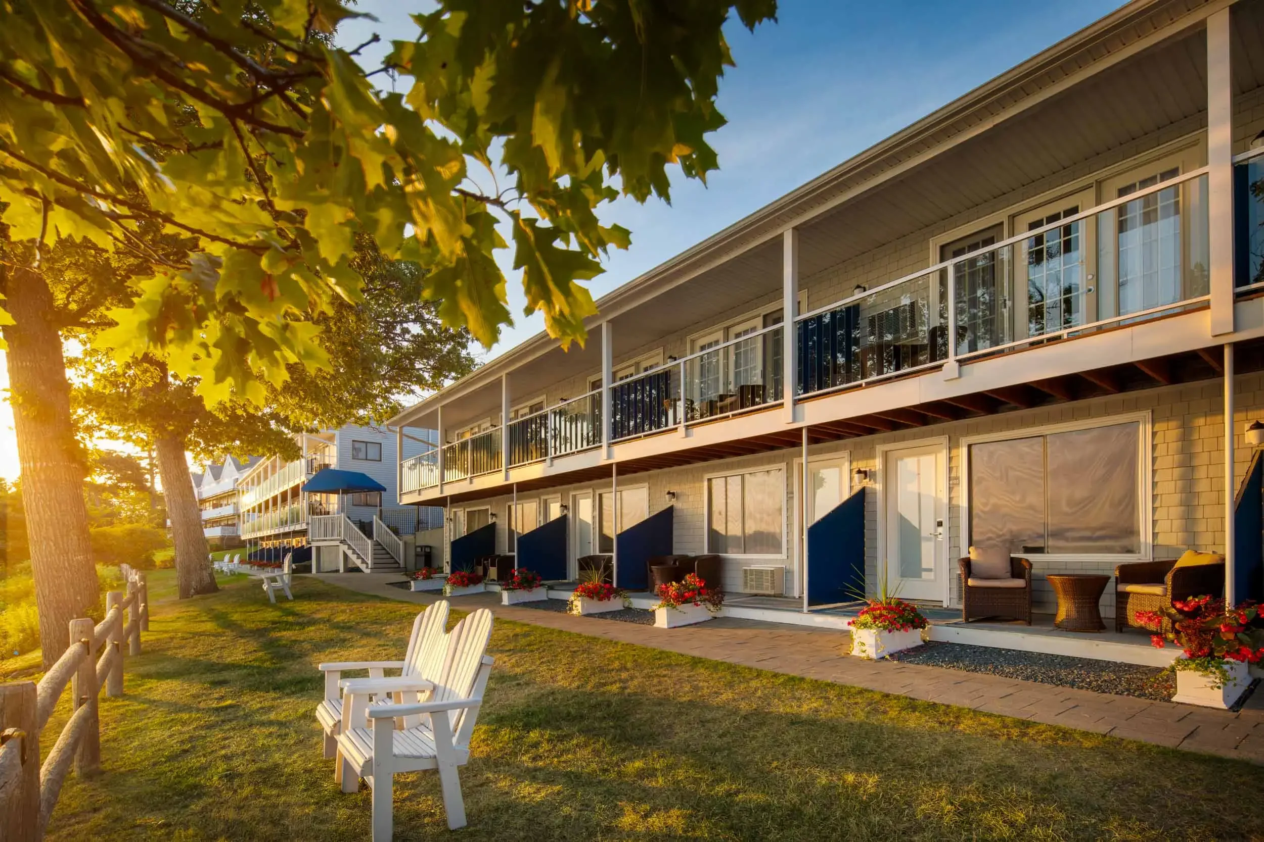 Sunset view of a multi-story residence with a balcony, patio chairs, and colorful flowers outside, surrounded by green trees on a grassy yard.