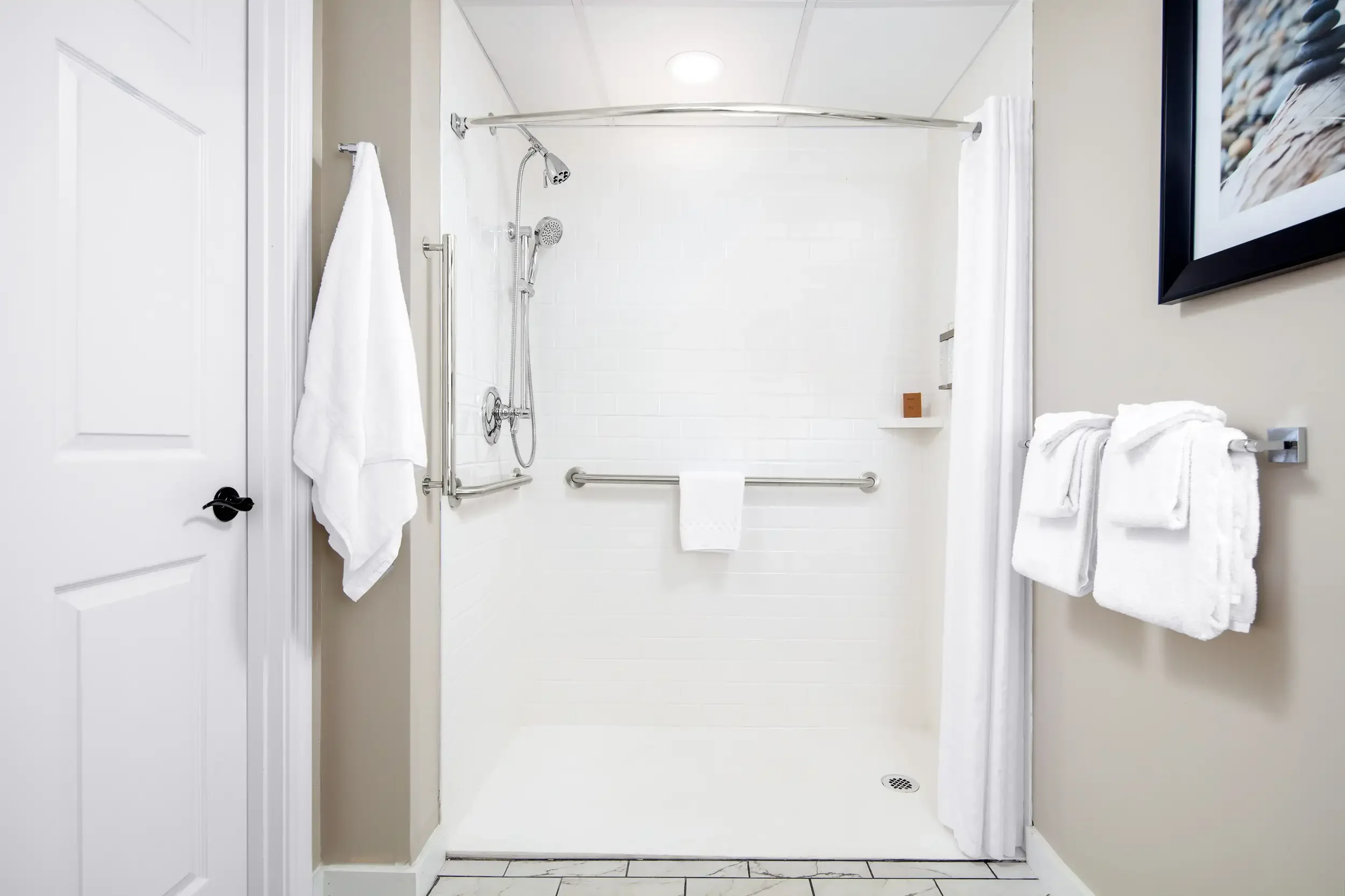 A walk-in shower with white tiled walls and a ceiling, featuring a grab bar, a hand-held showerhead, and a ceiling-mounted rain shower. There are white towels hanging, a small shelf with a soap or lotion bottle, and a framed picture partially visible