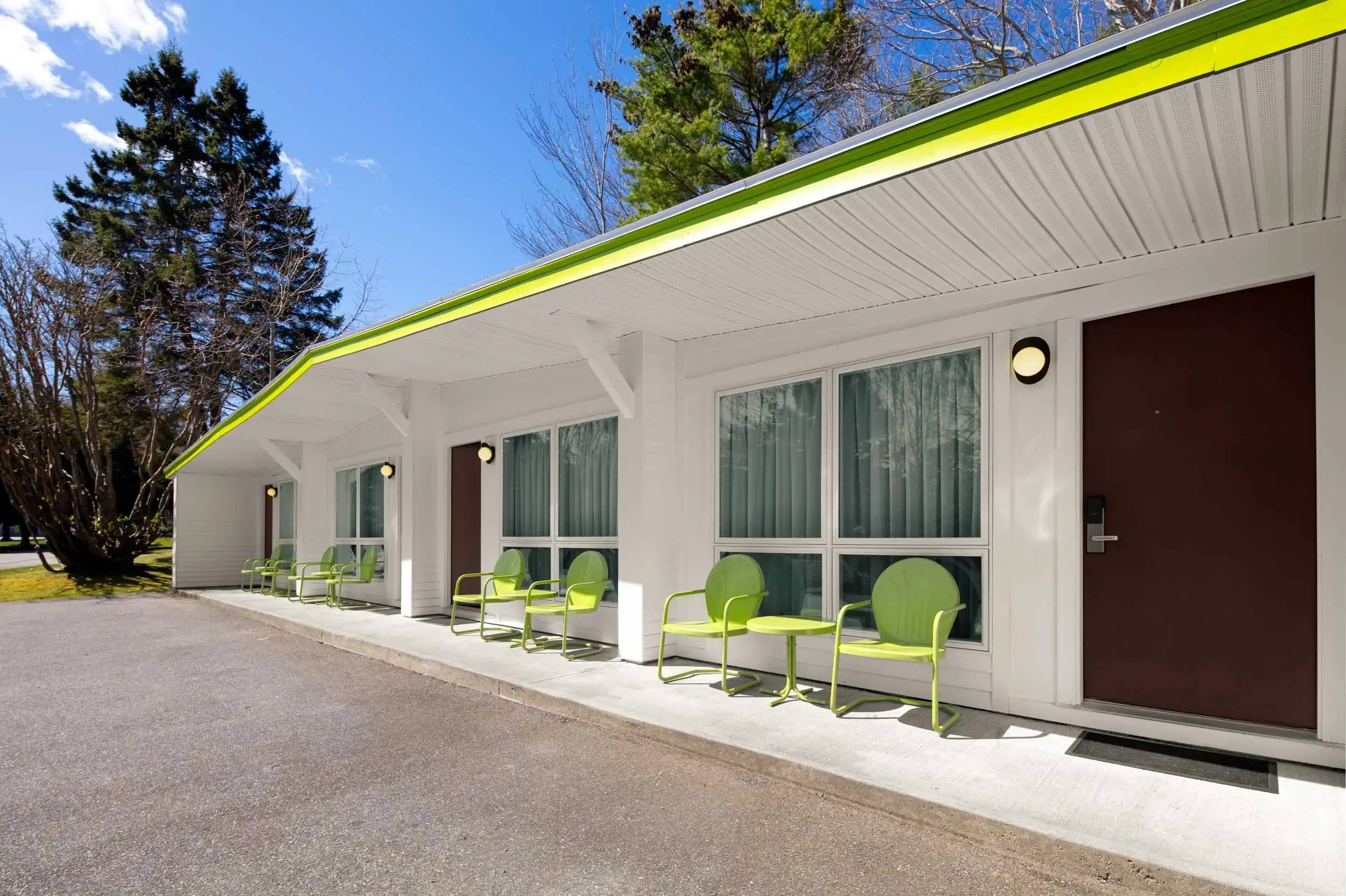 A row of lime green chairs outside a white building with large windows, a brown door, and a covered walkway with a sloped roof edged with lime green trim. There are trees in the background and a partly cloudy sky.