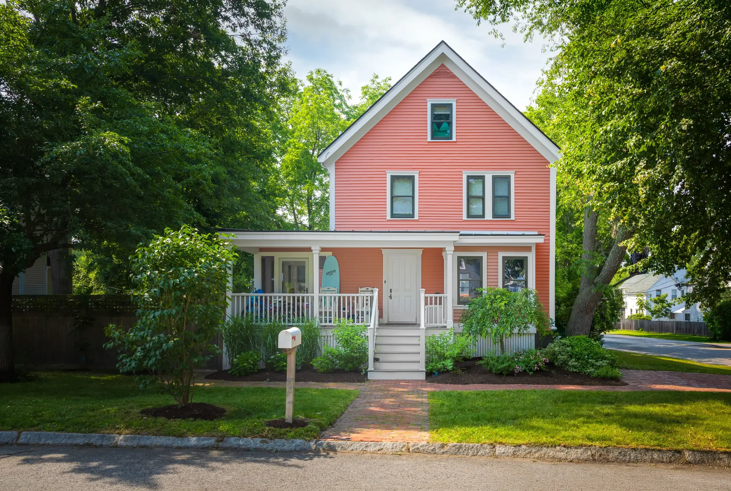 A pink two-story house with white trim and a small front porch, surrounded by green trees and a well-maintained lawn, with a brick pathway leading to the front door.
