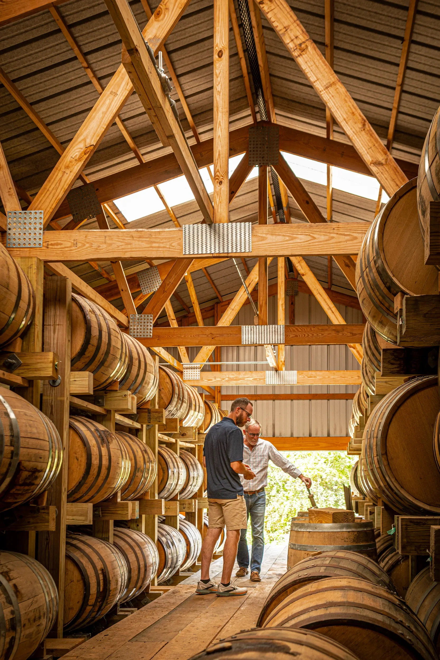 Two men standing inside a warehouse with wooden barrels on shelves, inspecting the barrels.