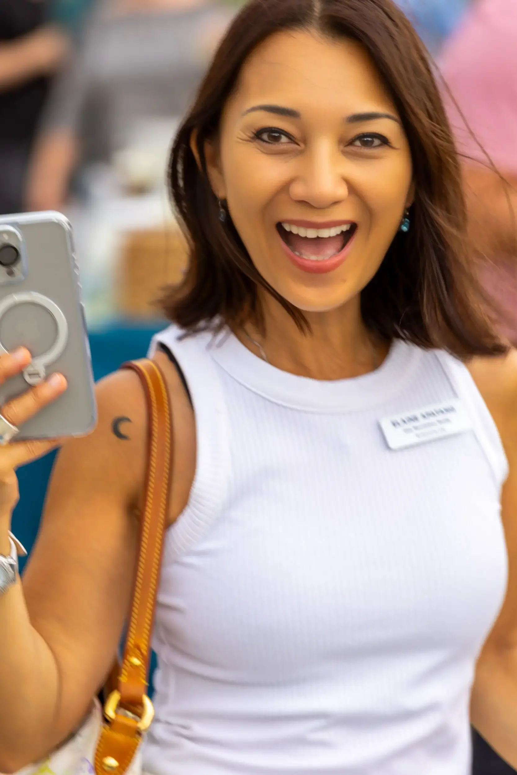 Close-up of a woman with brown hair taking a selfie, smiling, wearing a white sleeveless top, earrings, and carrying a brown shoulder bag. She has a tattoo on her upper arm.