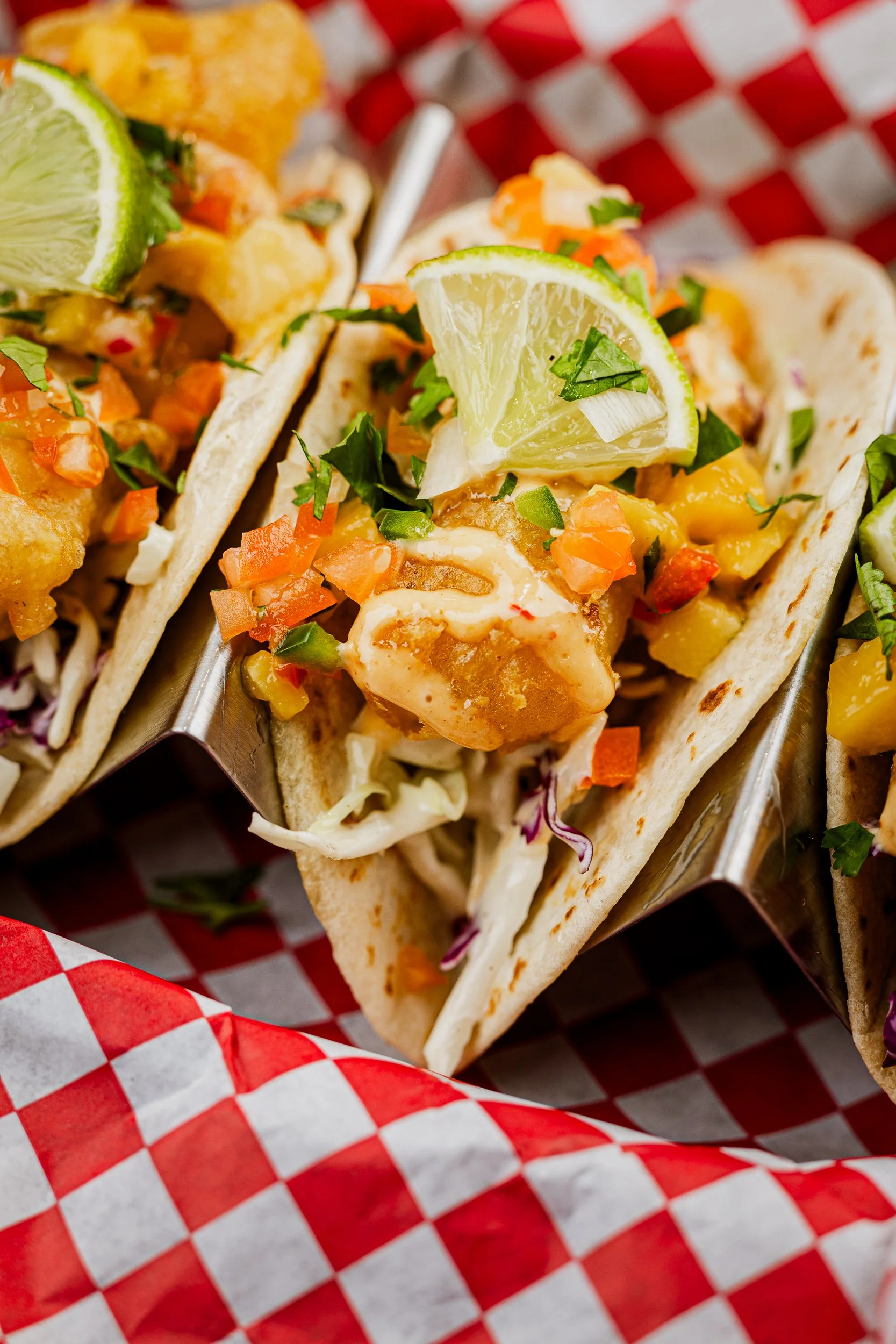 Close-up of fish tacos with fish, shredded cabbage, diced tomatoes, chopped cilantro, lime wedge, and creamy sauce in soft corn tortillas on red checkered paper.