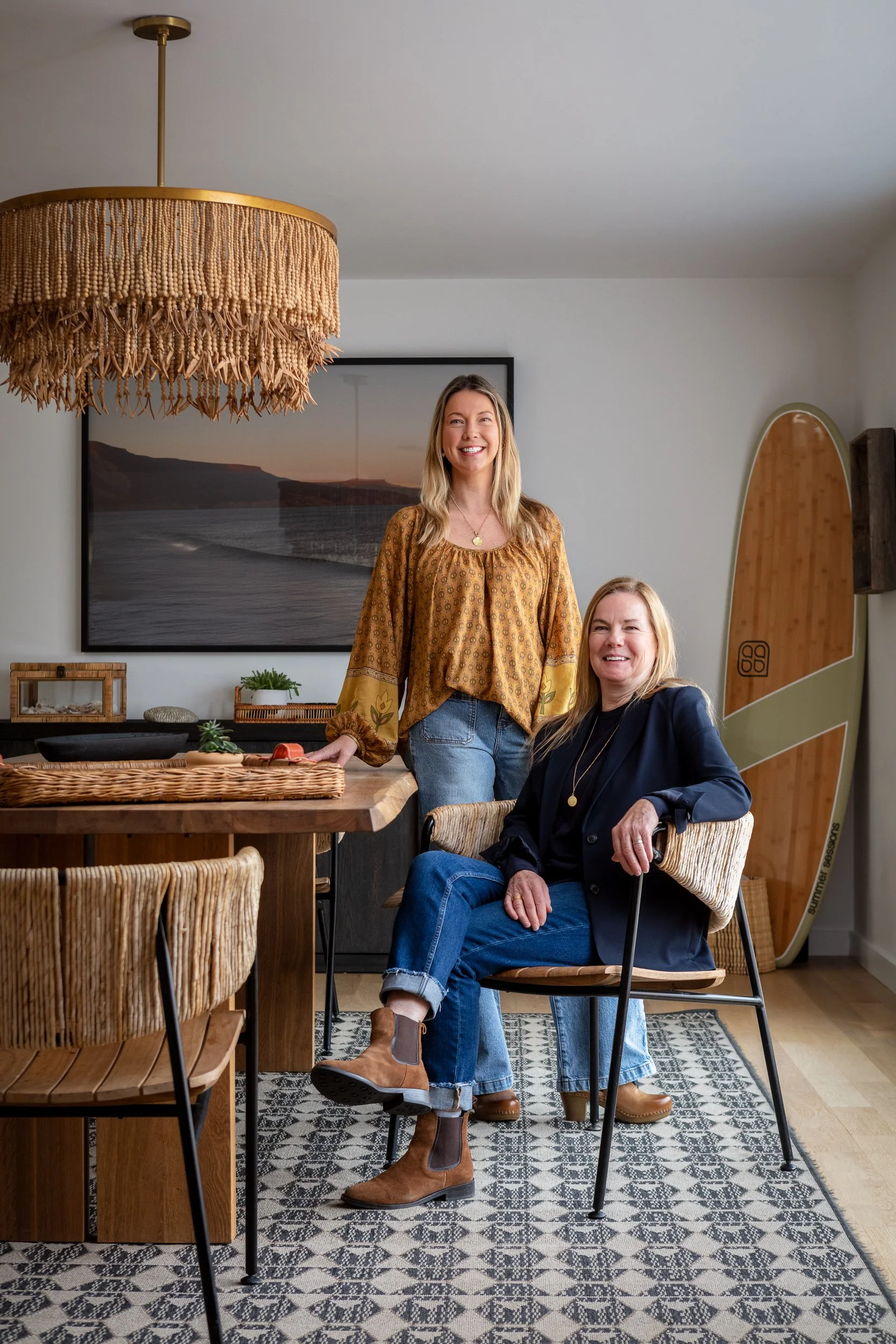 Two women in a dining room, one standing and the other sitting, smiling. The room has a wooden table, wicker chairs, a large framed sunset landscape on the wall, and a paddleboard leaning against the wall.