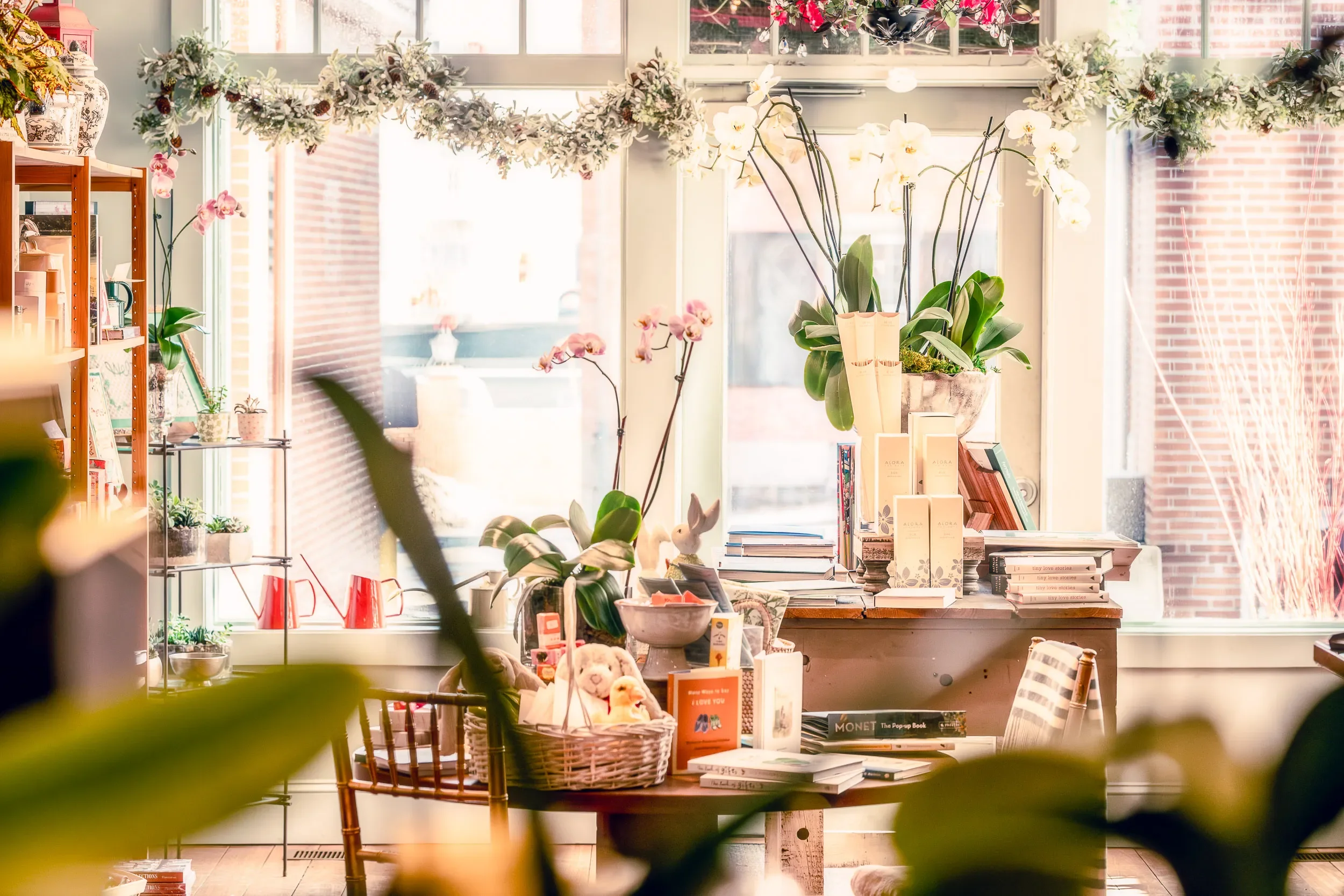 Bright, cozy interior of a floral shop or boutique with large windows, decorated with white orchids and pink flowers, and shelves filled with books, plants, and decor items.