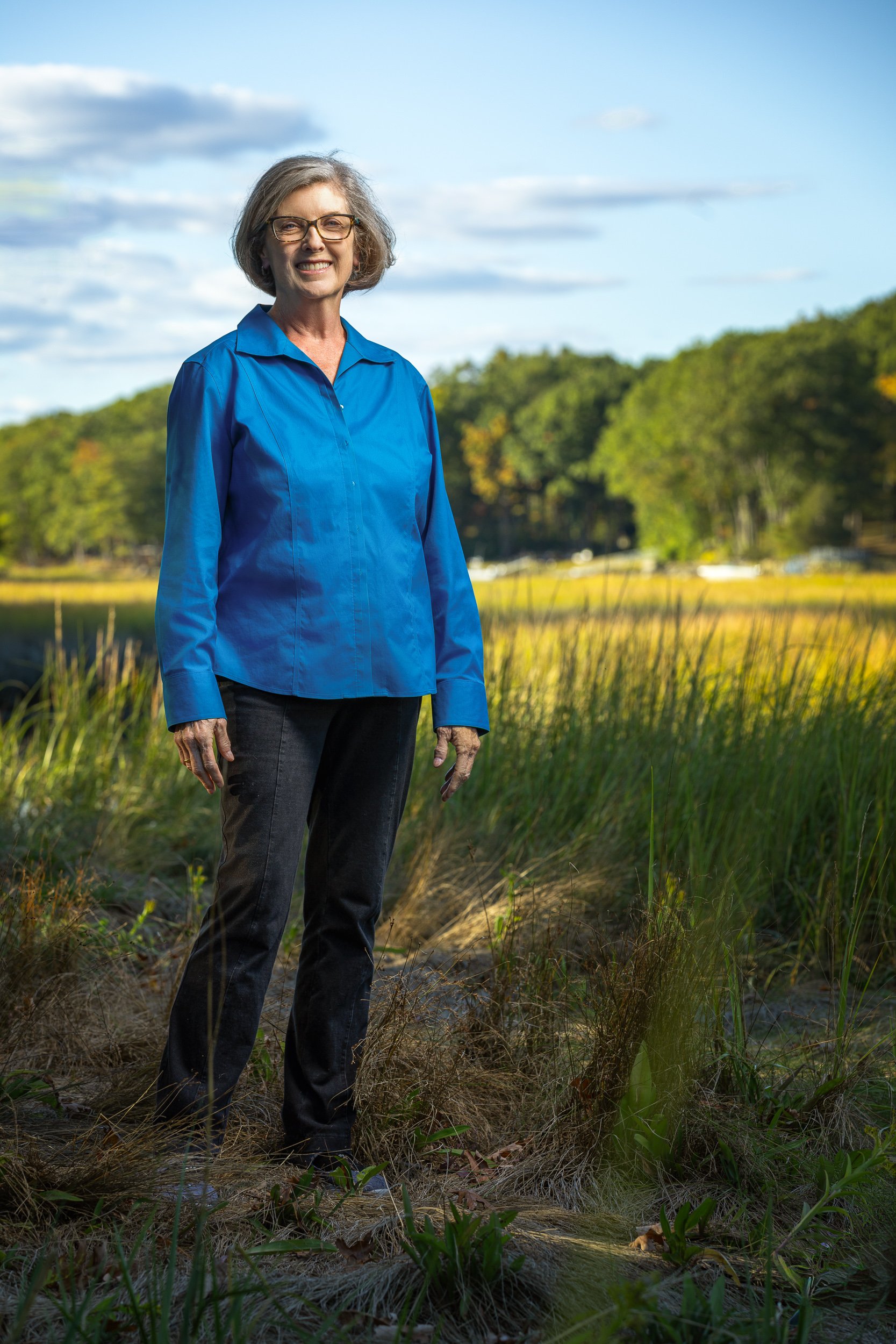A woman standing outdoors in a natural setting with trees and a body of water in the background, wearing a blue jacket and black pants, smiling.