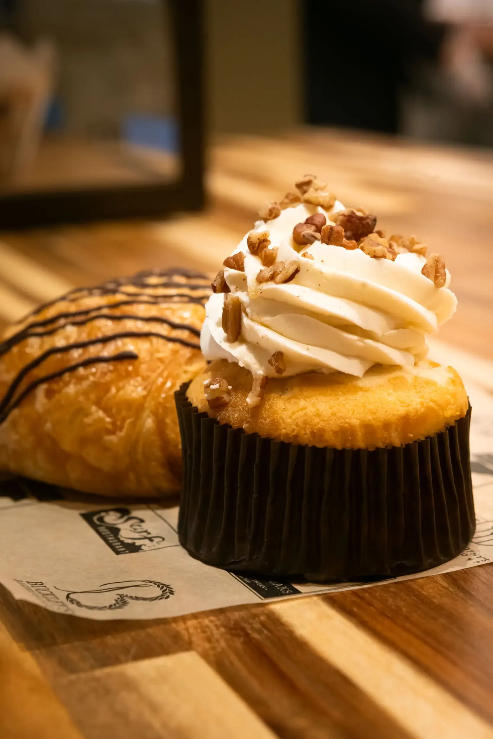 Cupcake with white frosting topped with chopped nuts, placed on a black cupcake liner on a piece of printed paper on a wooden table.