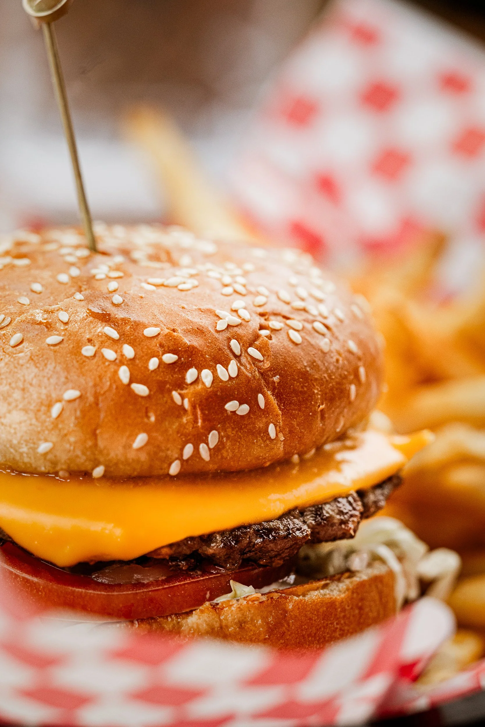 Close-up of a cheeseburger with a sesame seed bun, cheddar cheese, a beef patty, tomato, and lettuce, served with French fries in a basket.