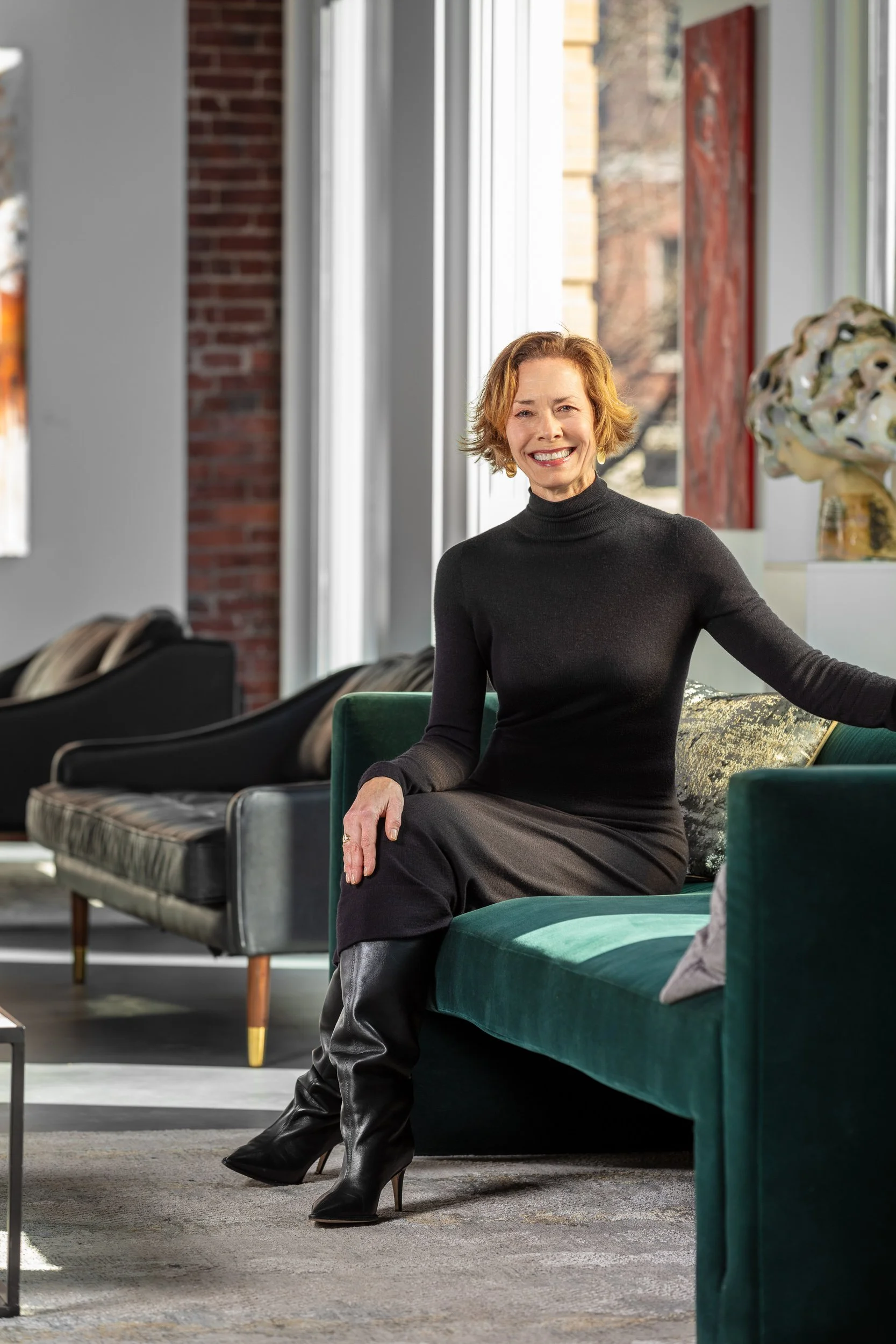 A woman with short red hair, wearing a black turtleneck and black boots, is sitting on a green velvet couch, smiling in a brightly lit room with large windows and colorful artwork.