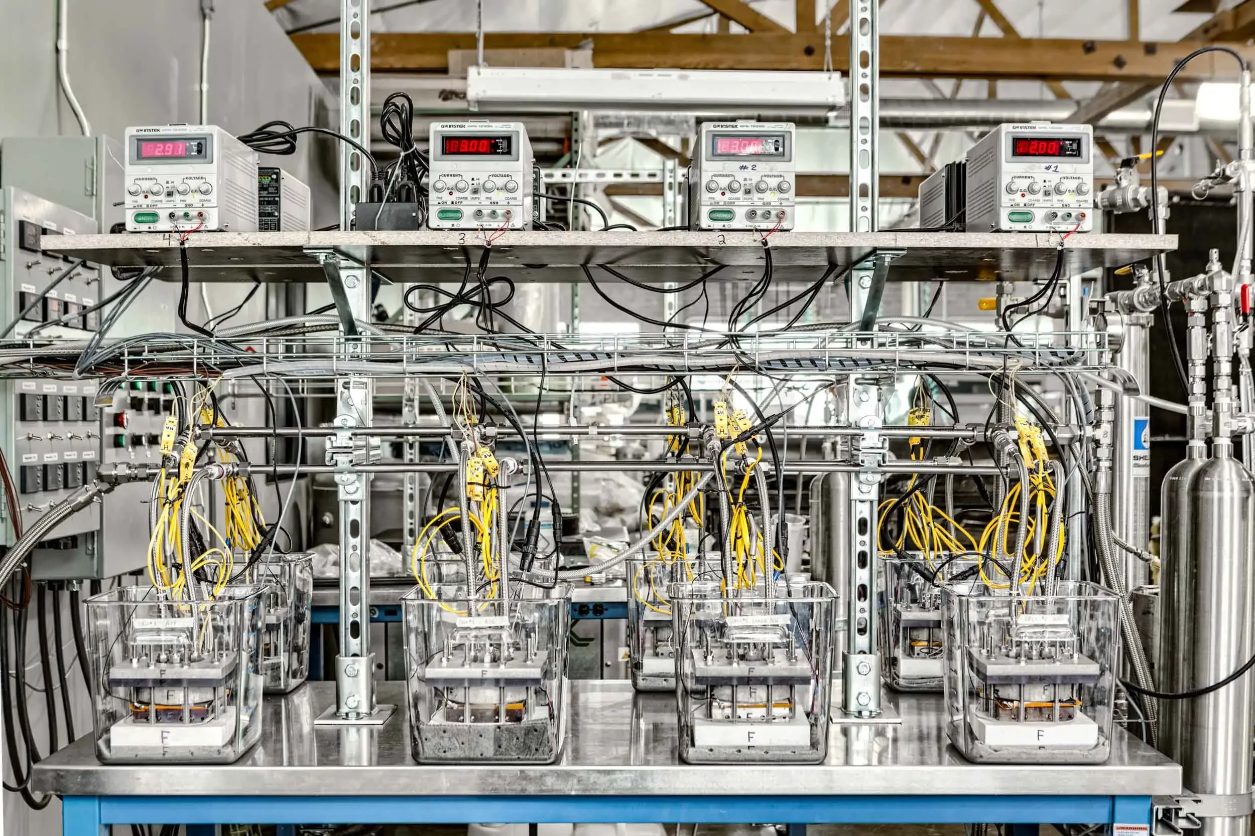 Laboratory setup with multiple transparent containers connected by numerous yellow and black wires, electronic equipment with digital displays, and stainless steel plumbing components.