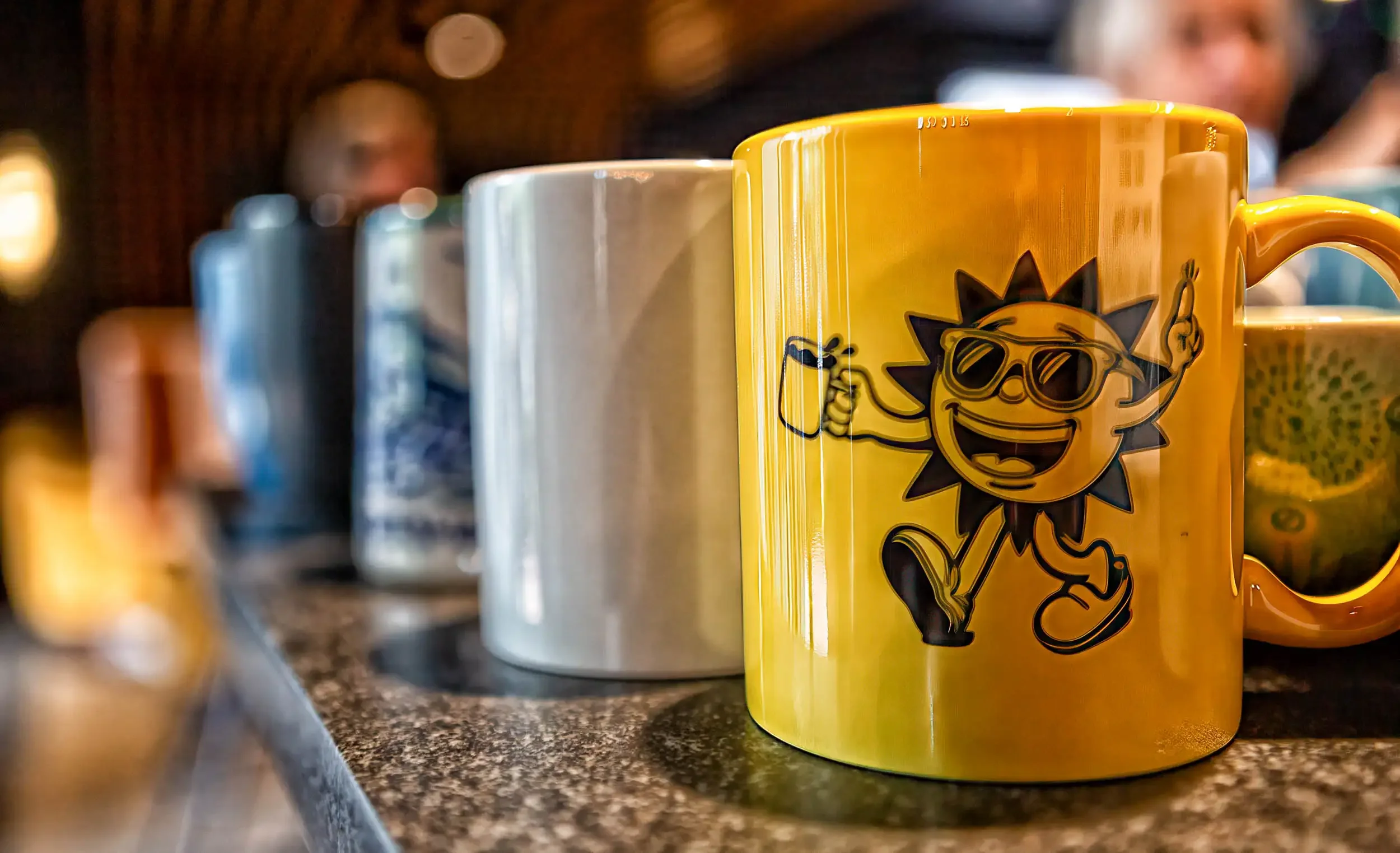 Close-up of a yellow coffee mug with a cartoon lion wearing sunglasses printed on it, placed on a dark granite countertop alongside other mugs, with a blurred background of people in a cafe.