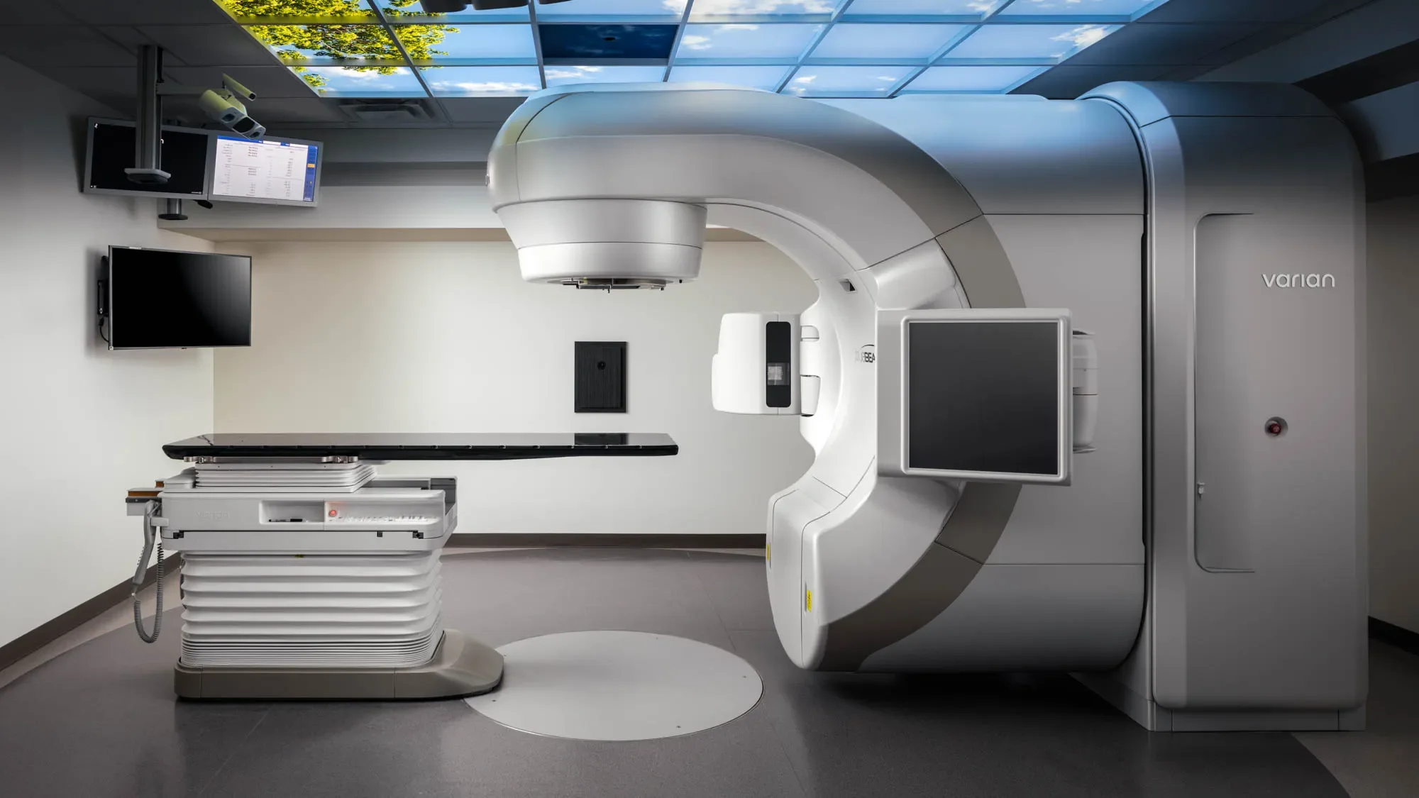Medical imaging room with a large MRI machine labeled 'varian' and multiple monitors on the wall.