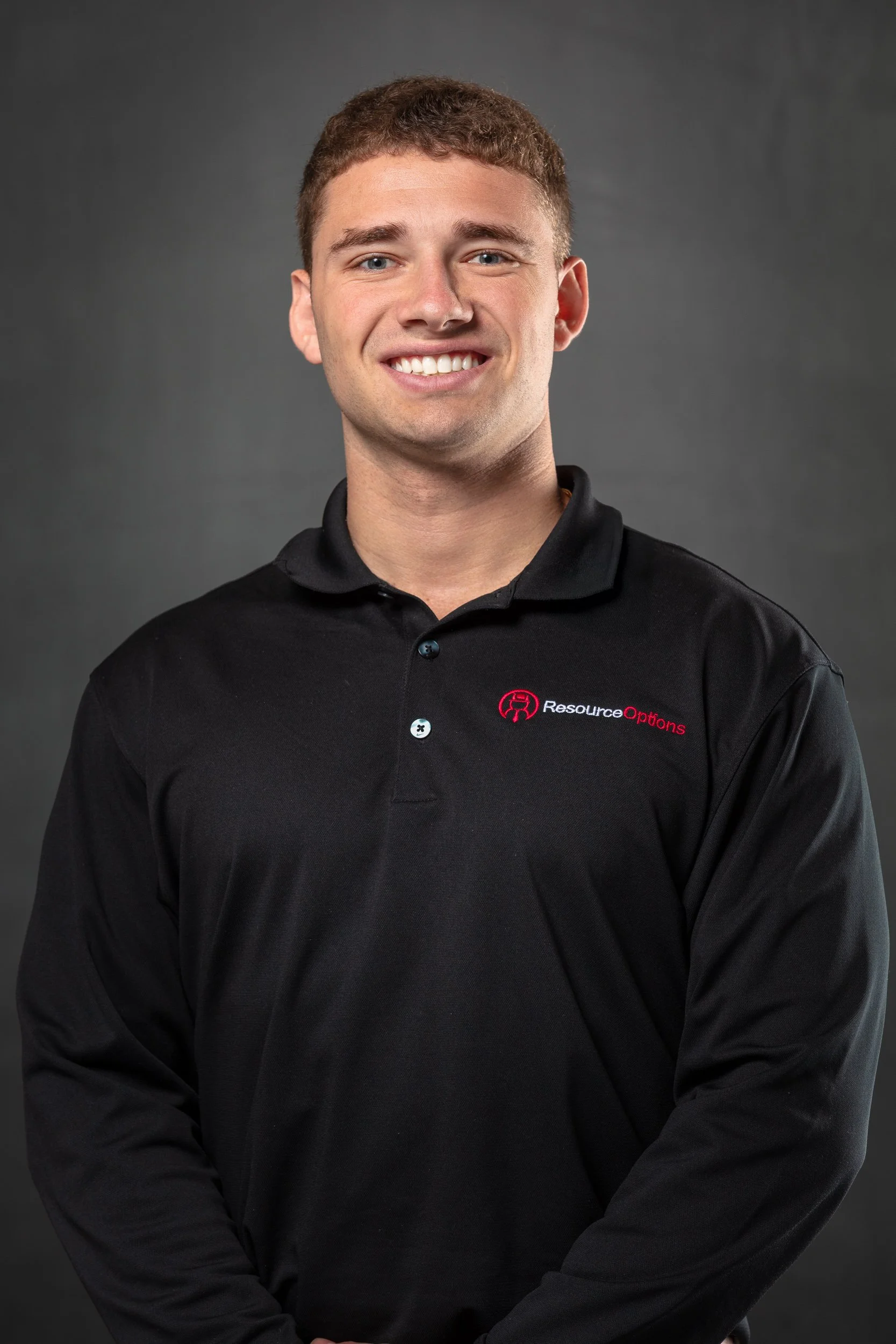 A young man with short brown hair wearing a black polo shirt with a logo that reads 'Resource Options'. He is smiling and standing against a plain dark gray background.
