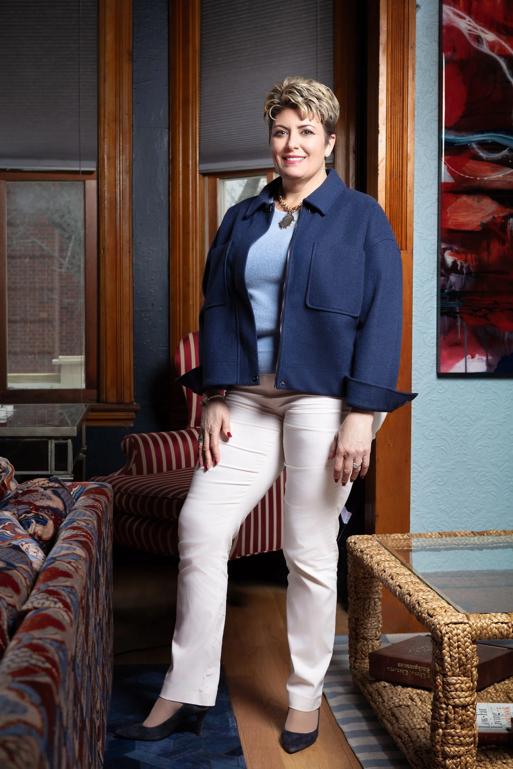 A woman with short, light brown hair standing in a room with wooden window frames, wearing a navy blue jacket, light blue sweater, white pants, black high heels, and a statement necklace. She is smiling and standing next to a patterned armchair and a