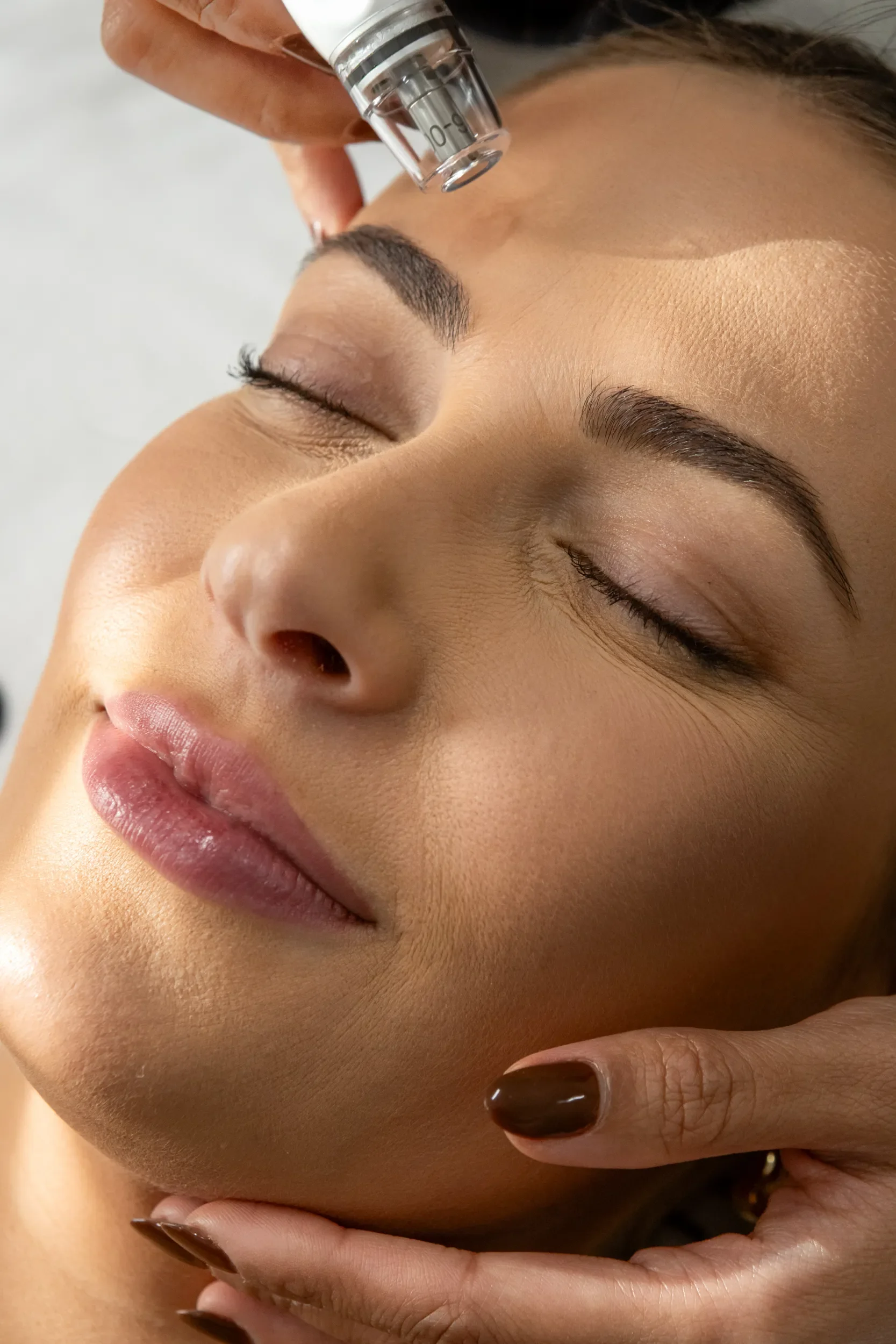 A person receiving a cosmetic facial treatment, with a close-up of their face showing closed eyes and smooth skin. A practitioner's hand holds a small tool or device near the person's forehead.