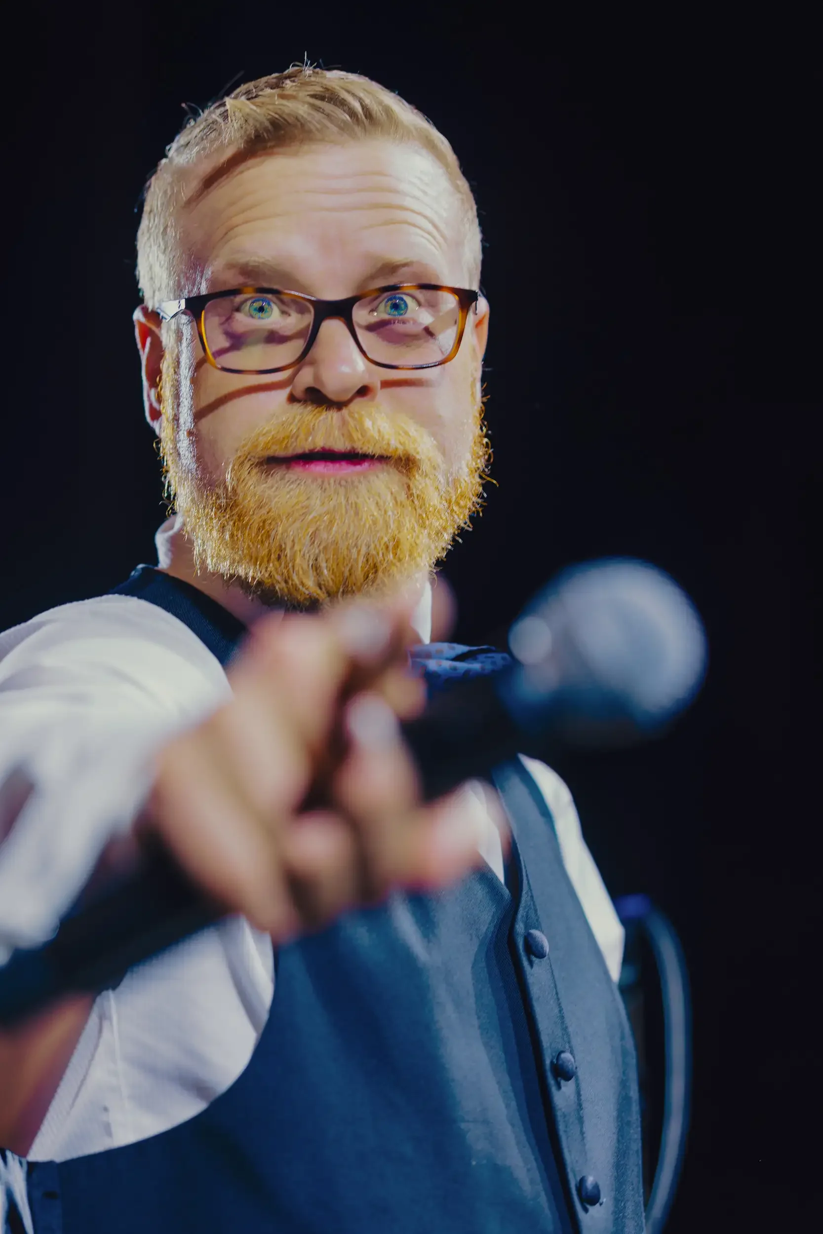 A man with glasses, a ginger beard, and hair pointing towards the camera in front of a microphone, wearing a vest and a white shirt, with a black background.
