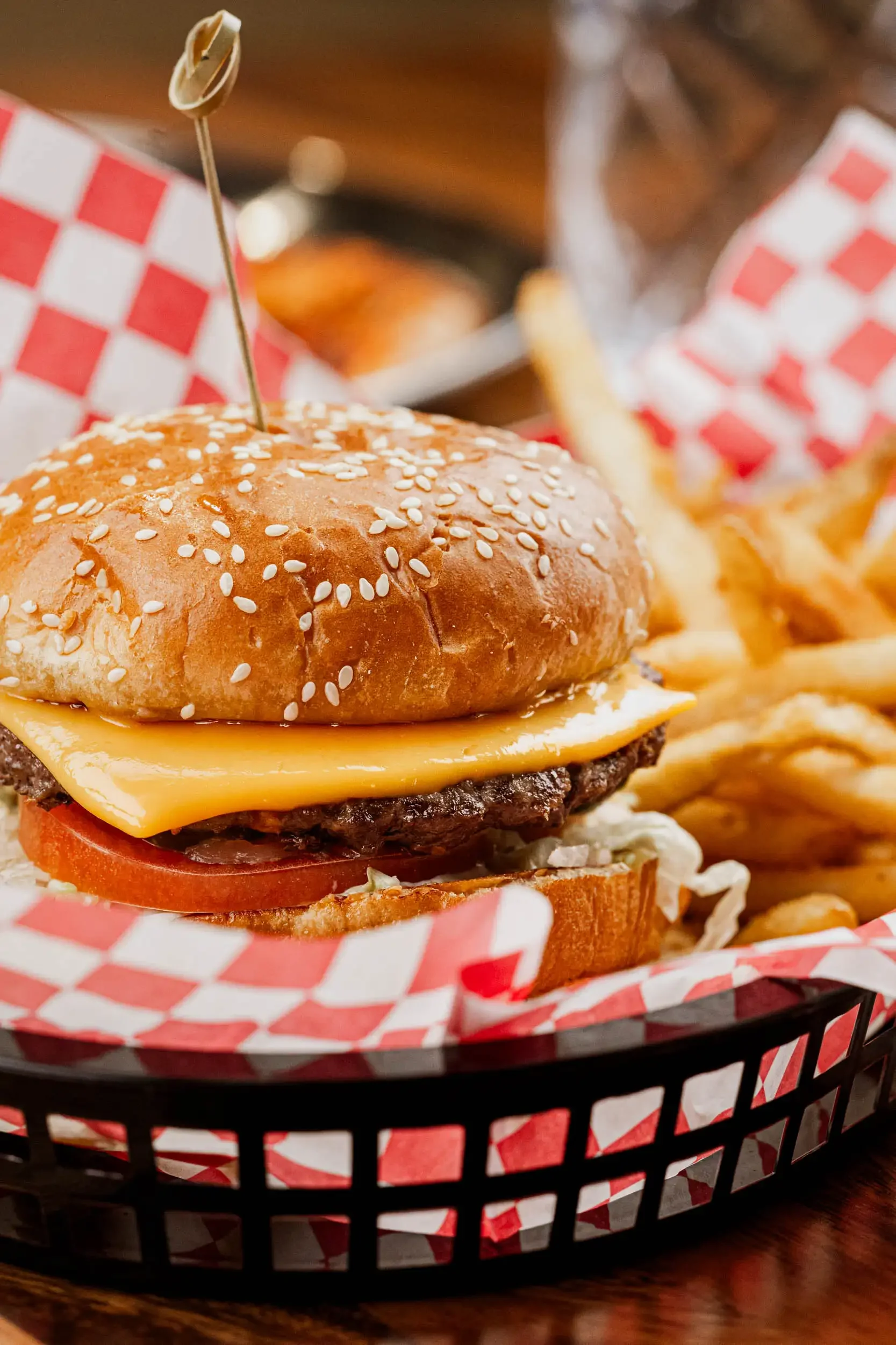 Close-up of a cheeseburger with a sesame seed bun, lettuce, tomato, cheese, and a beef patty, served in a black basket lined with red and white checkered paper, with fries in the background.
