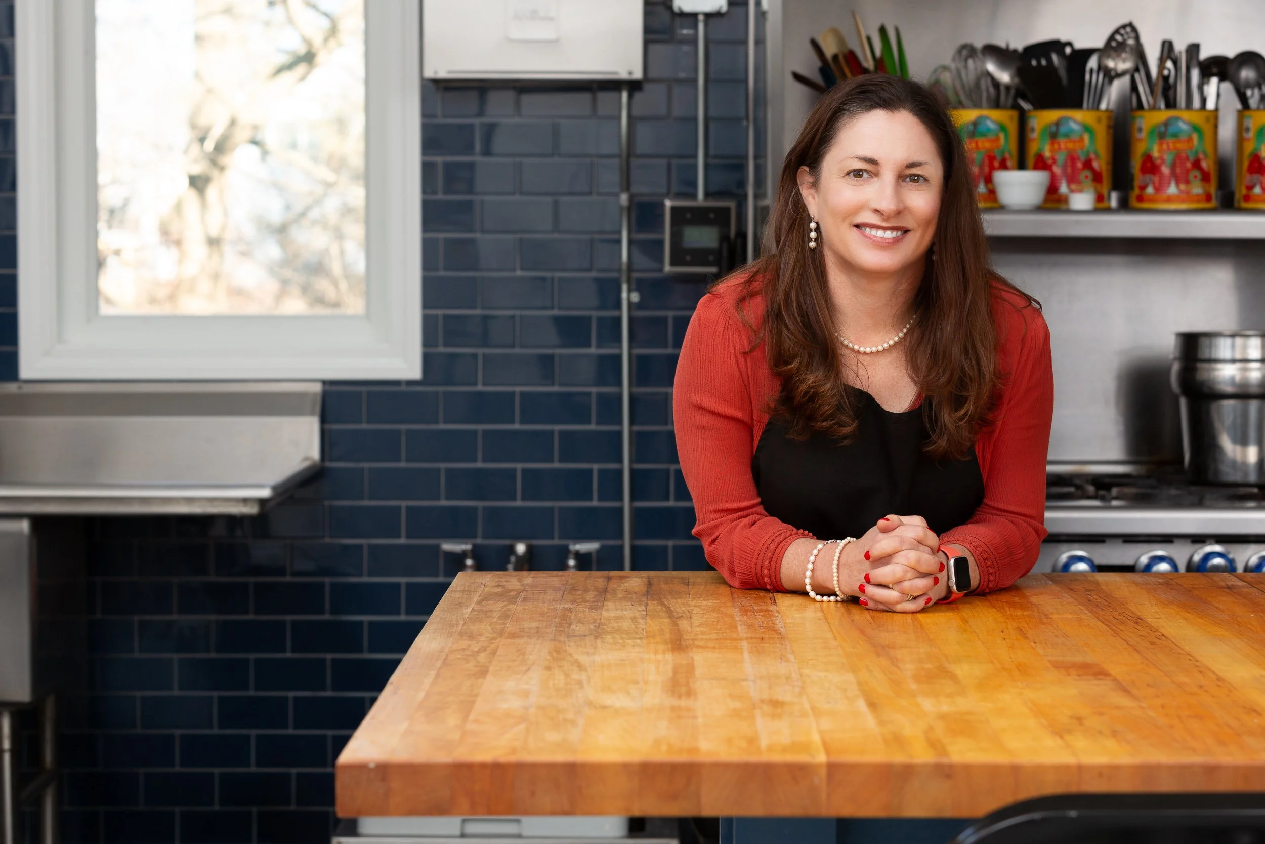 A woman with brown hair wearing a black top and red cardigan, accessorized with pearl jewelry, smiling and leaning on a wooden kitchen counter with her hands clasped together. The kitchen has dark blue tiles, a window, and shelves with canned goods a