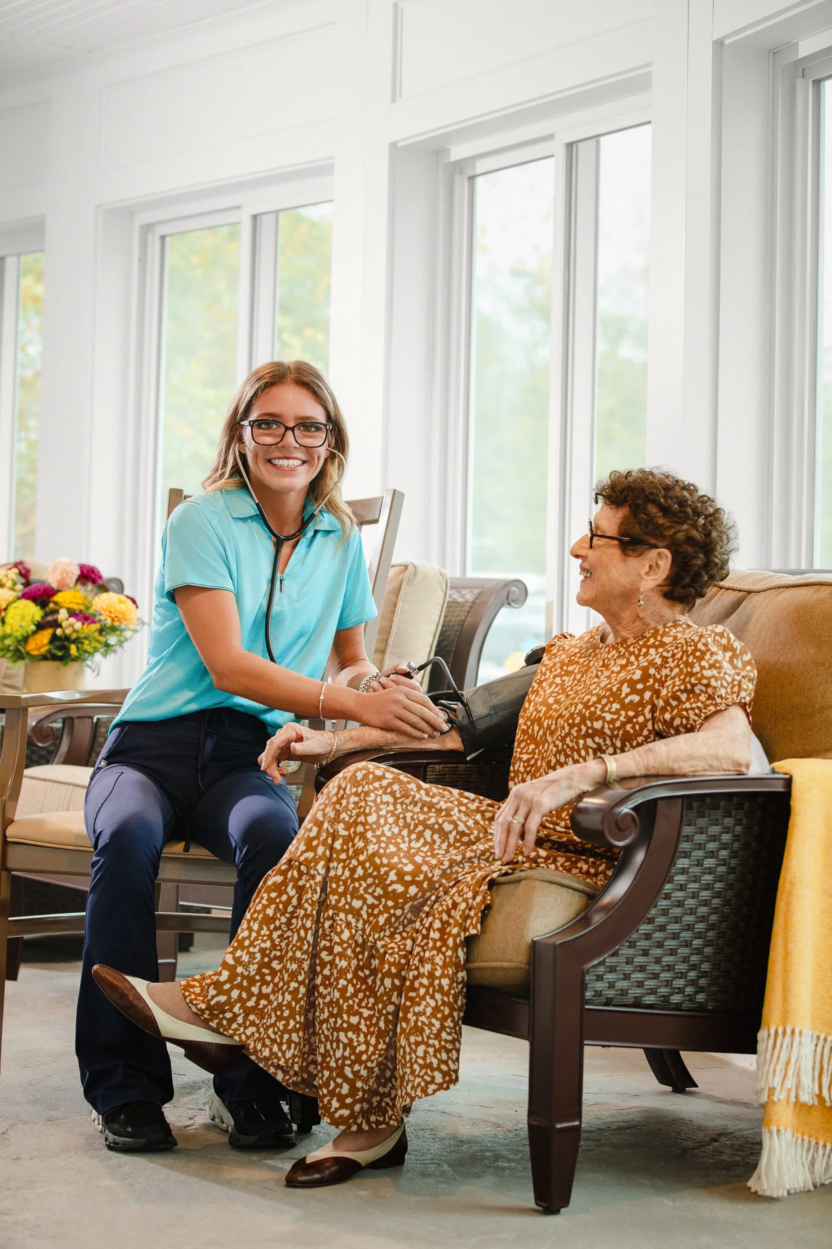 A healthcare professional, wearing a stethoscope and scrubs, checks the blood pressure of an elderly woman sitting on a couch in a bright living room.