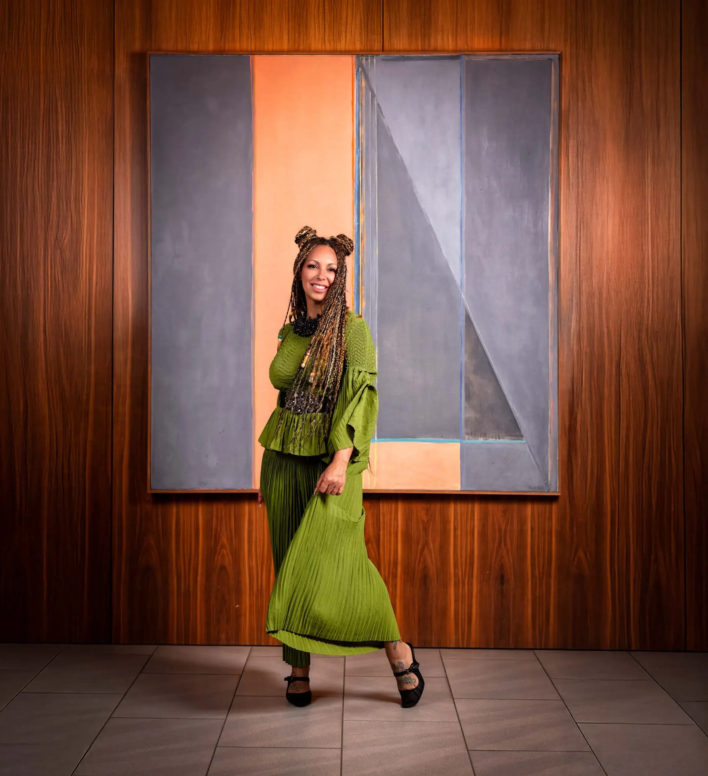 Woman standing in front of a wooden wall with colored panels, wearing a green pleated skirt, black high heels, and black jewelry, smiling with hair styled in braids in two buns.