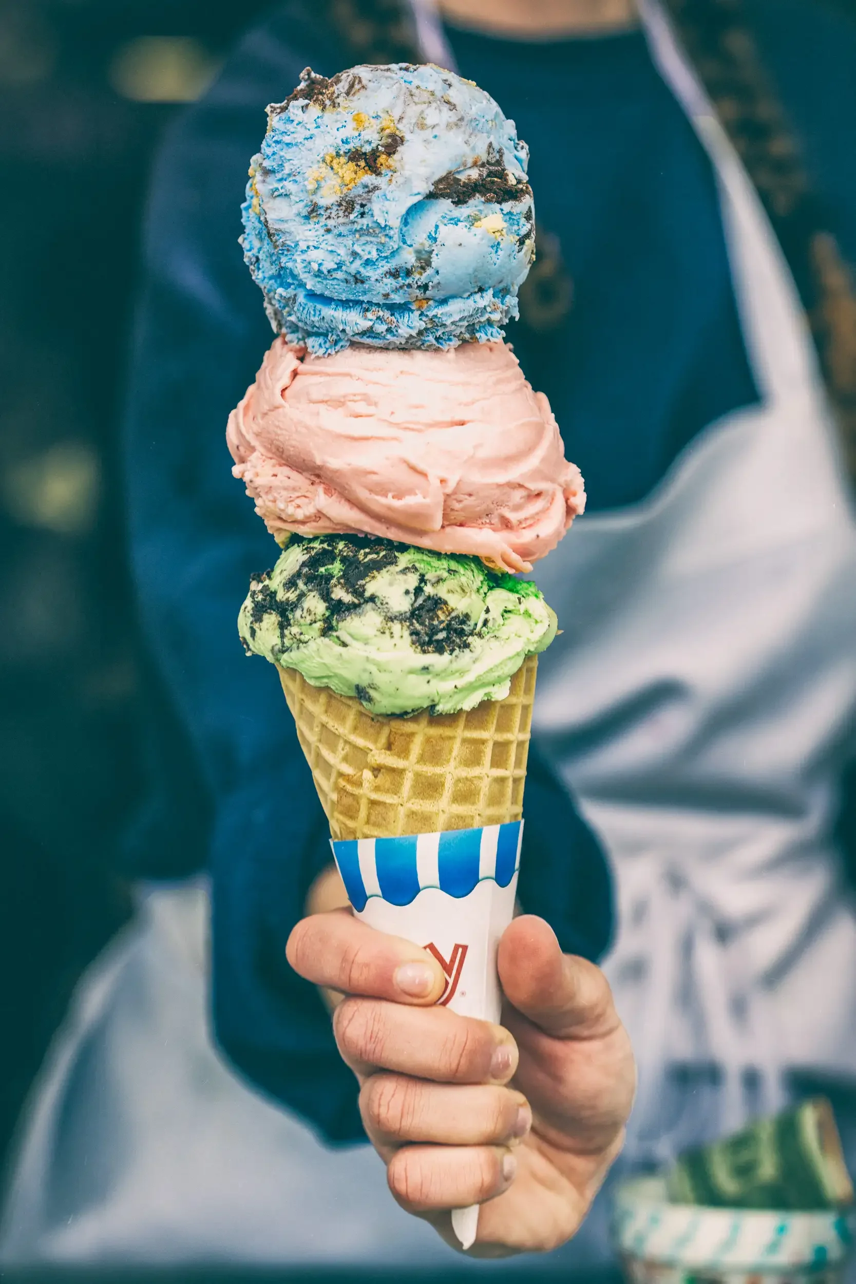 A hand holding a triple scoop ice cream cone with pink, blue, and green ice cream, in a waffle cone with a blue and white paper sleeve.