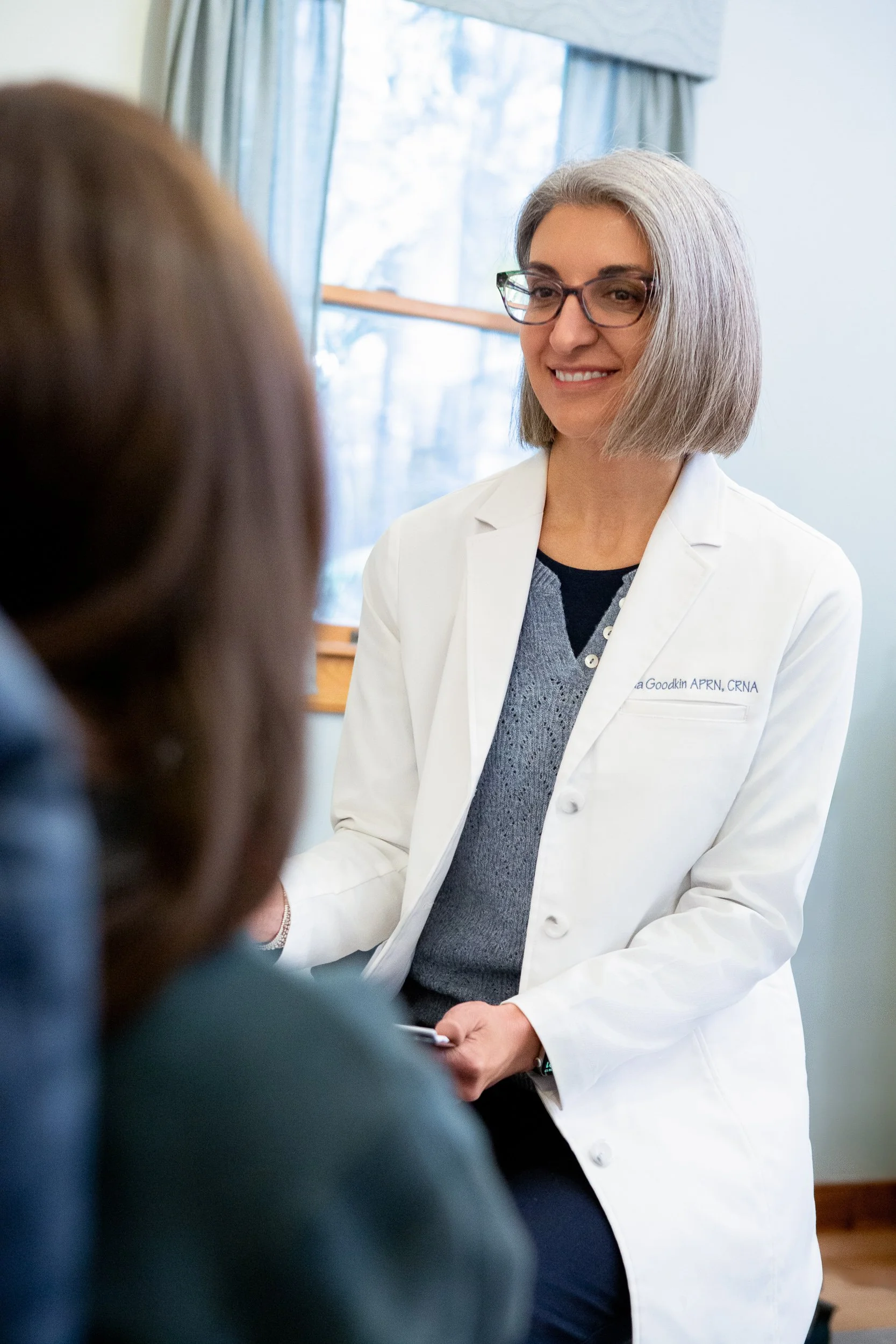 A female doctor with short gray hair and glasses, wearing a white coat, talking to a patient in an office with a window in the background.