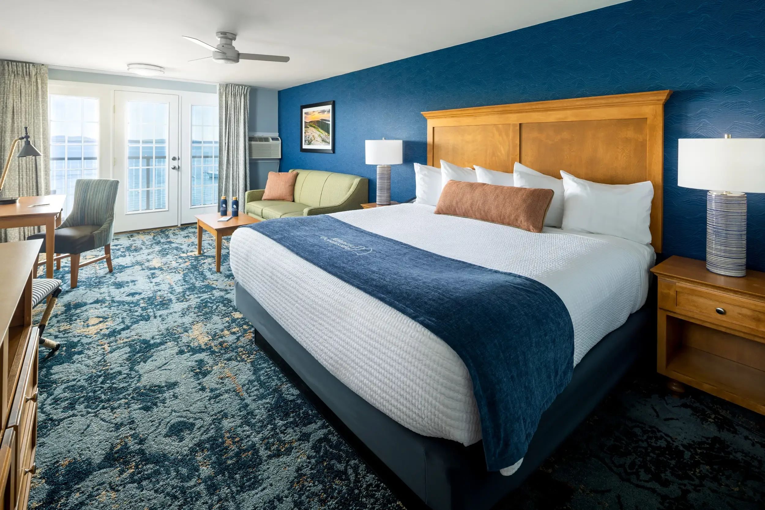 A hotel room with a large bed, wooden headboard, bedside lamps, couch, table, and a balcony with glass door, decorated with blue walls and patterned carpet.