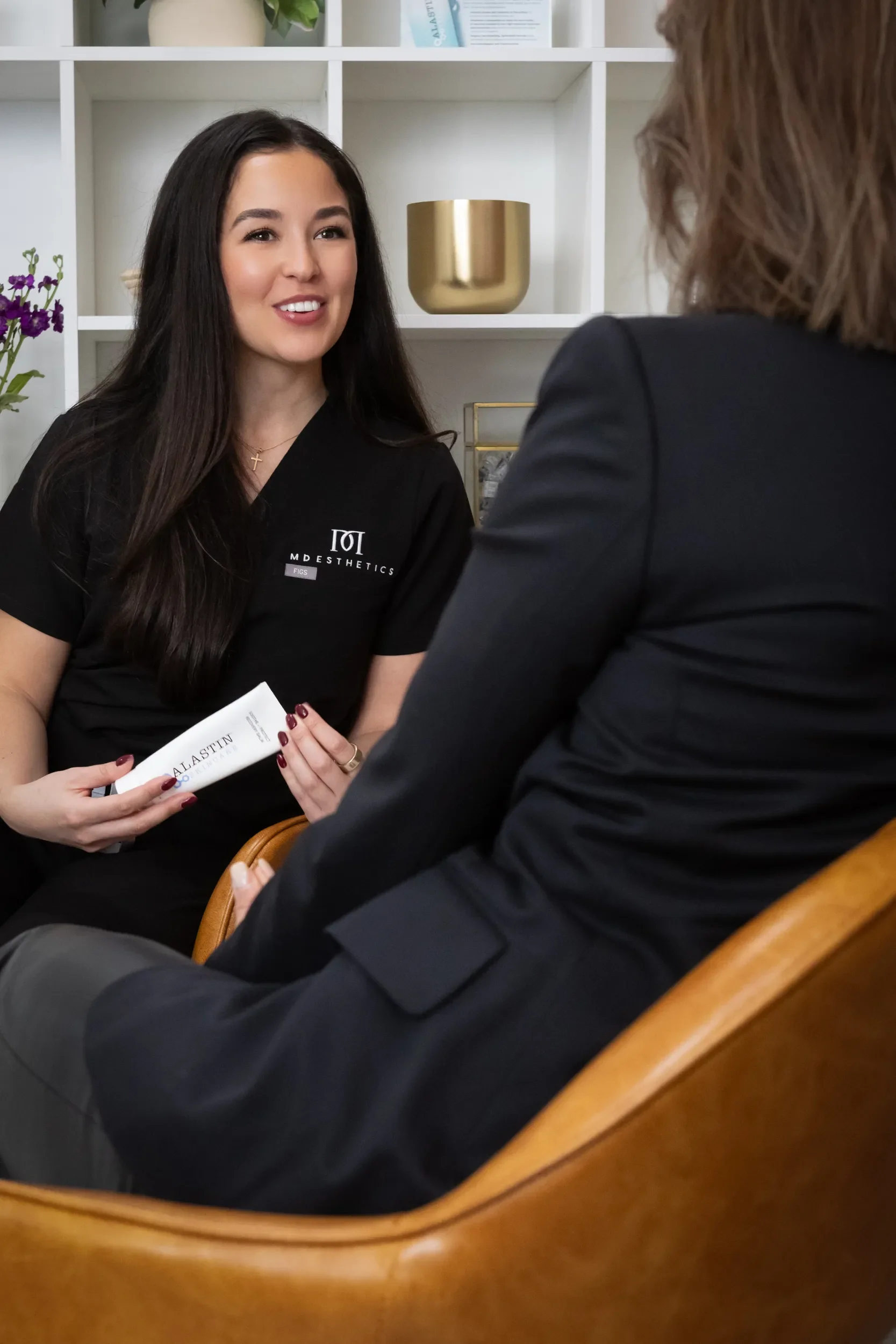 A woman with long dark hair, wearing a black shirt with a logo, is smiling while talking to another woman. The woman is holding a brochure. They are seated in a wooden chair in a room with a white bookshelf behind her, decorated with a gold pot and o