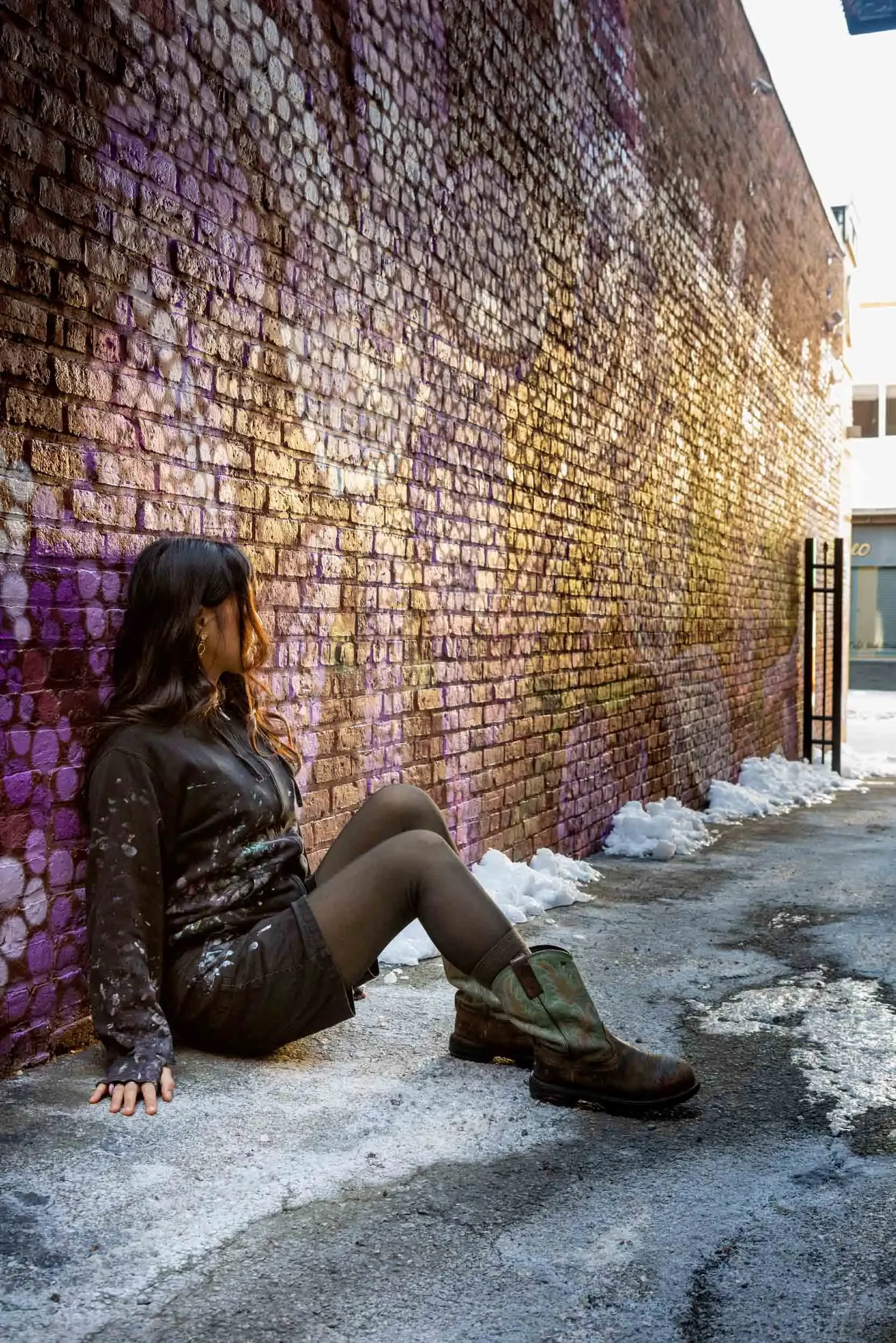 A woman with dark hair sitting on the ground leaning against a brick wall, wearing a dark jacket, shorts, and boots, with snow on the ground and a faint typo on the wall that spells 'TRUST.'