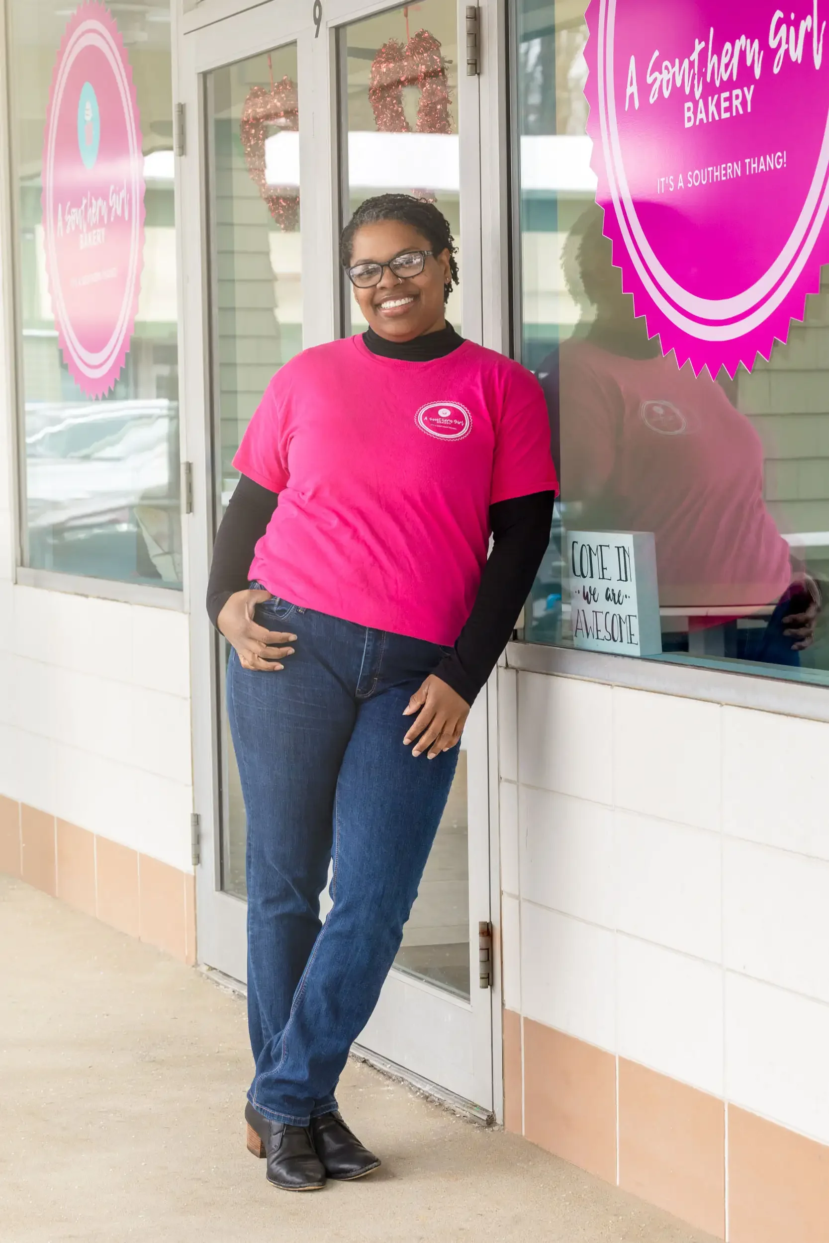 Woman standing outside a bakery, smiling, wearing glasses, a pink shirt with bakery logo, jeans, and black boots, in front of glass doors and pink signage.