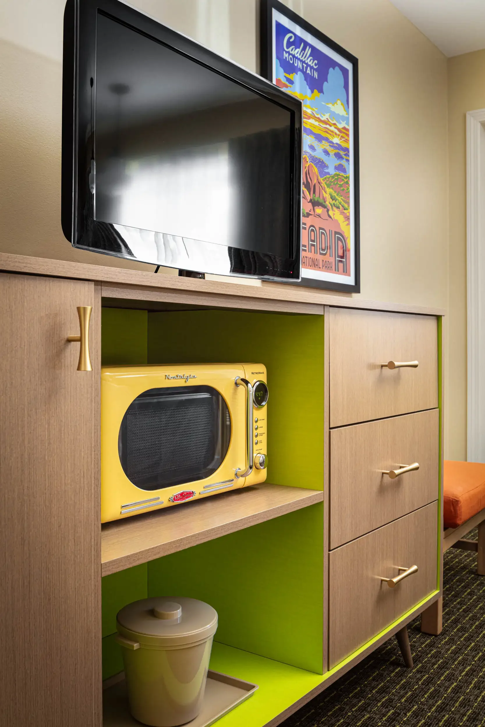 A wooden shelving unit with green back panels holds a yellow vintage toaster oven, a small white container, and a white trash bin. A black television is mounted above the unit, and a colorful poster of Cadillac Mountain is hanging on the wall nearby.
