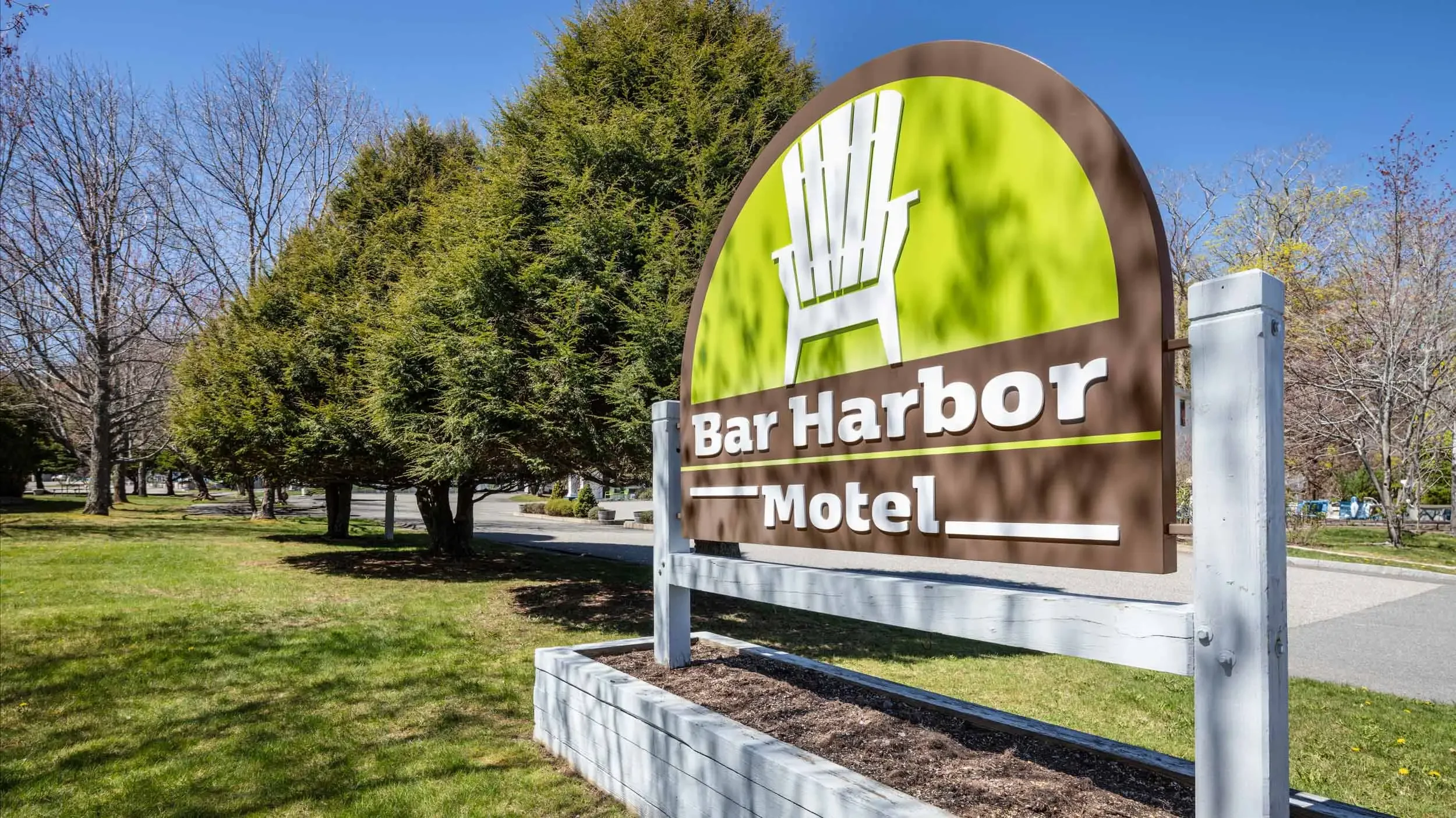 Sign for Bar Harbor Motel with a green background, stylized palm tree logo, located in a park-like setting with trees and a grassy area.