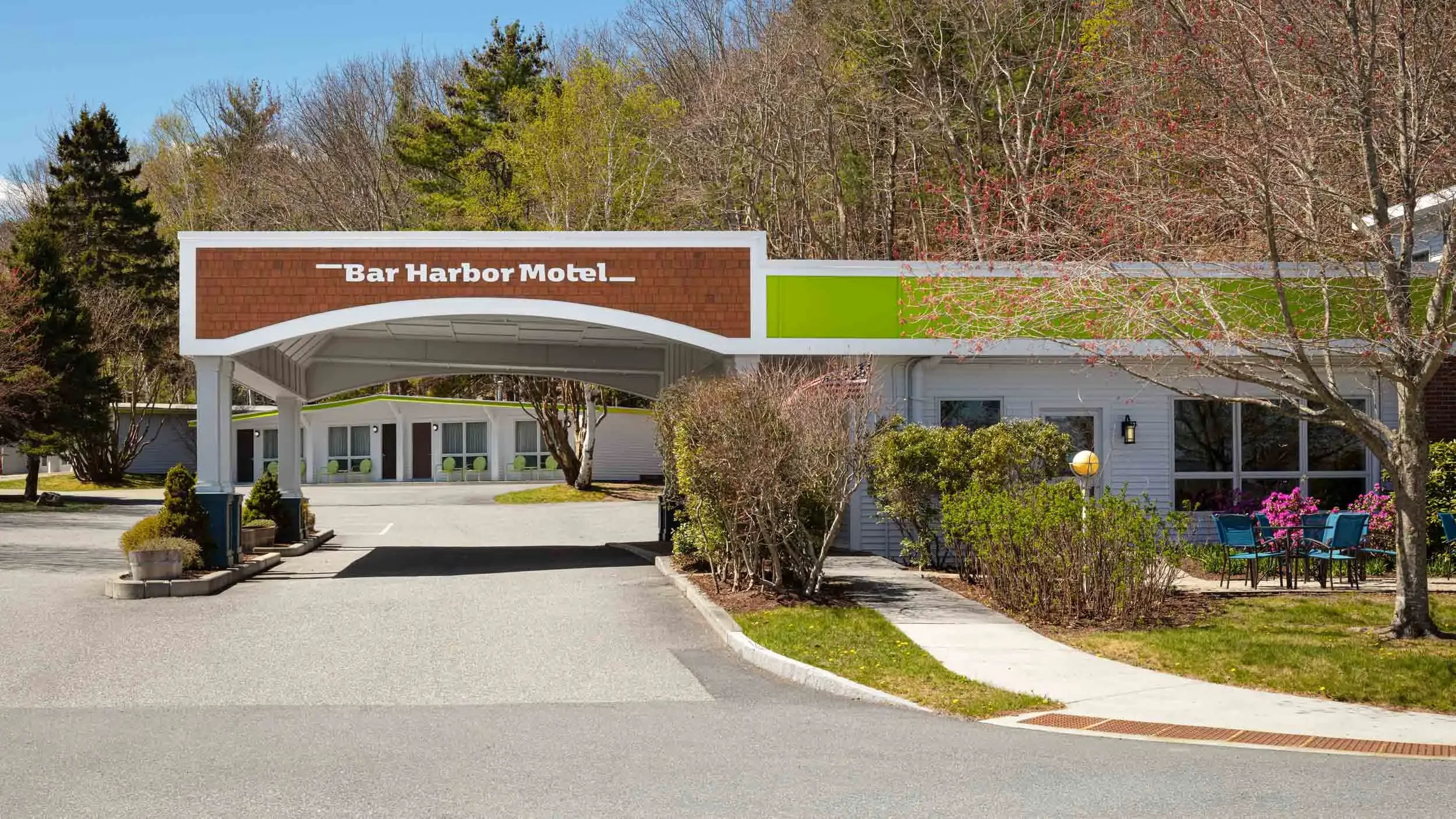 Exterior view of the Bar Harbor Motel with a parking lot, trees, bushes, and outdoor seating area, under a clear blue sky.