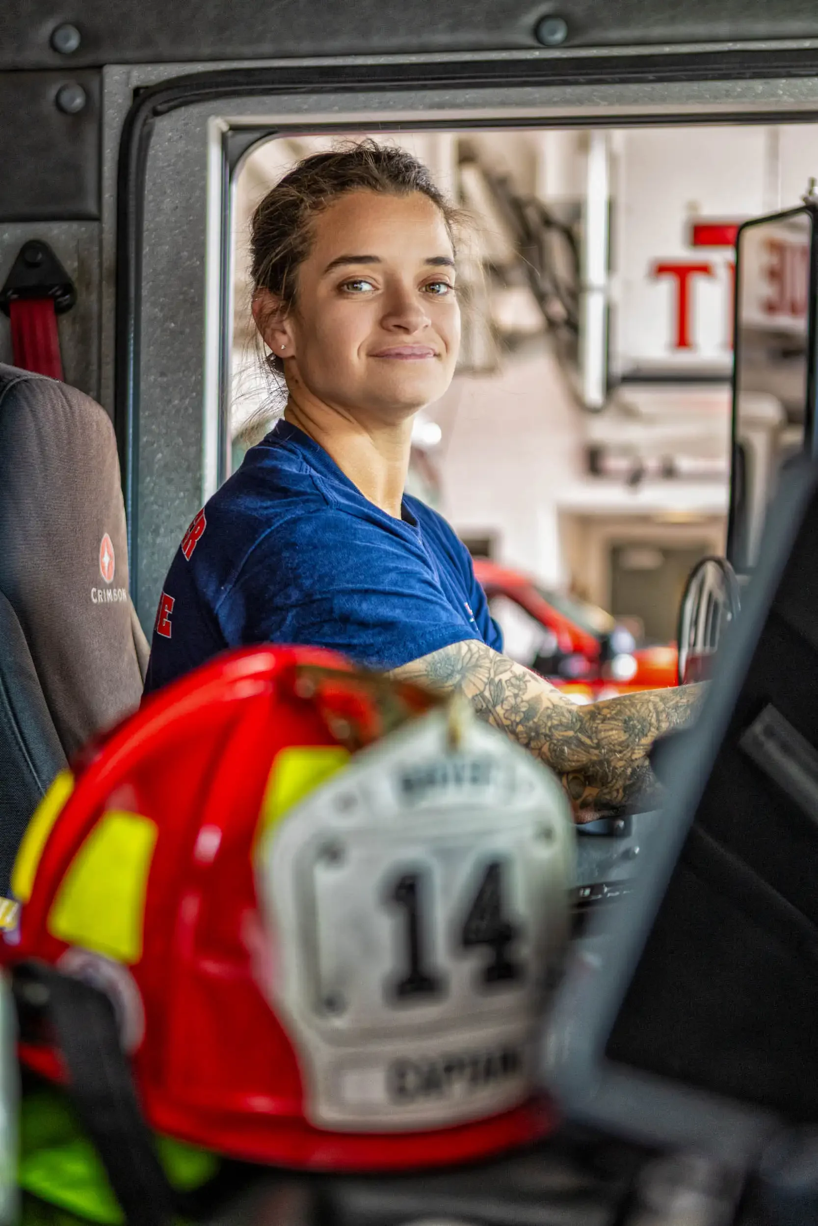 A female firefighter with blonde hair pulled back, wearing a blue uniform, sitting inside a fire truck, looking out the window, with firefighting equipment and a red fire helmet in the foreground.