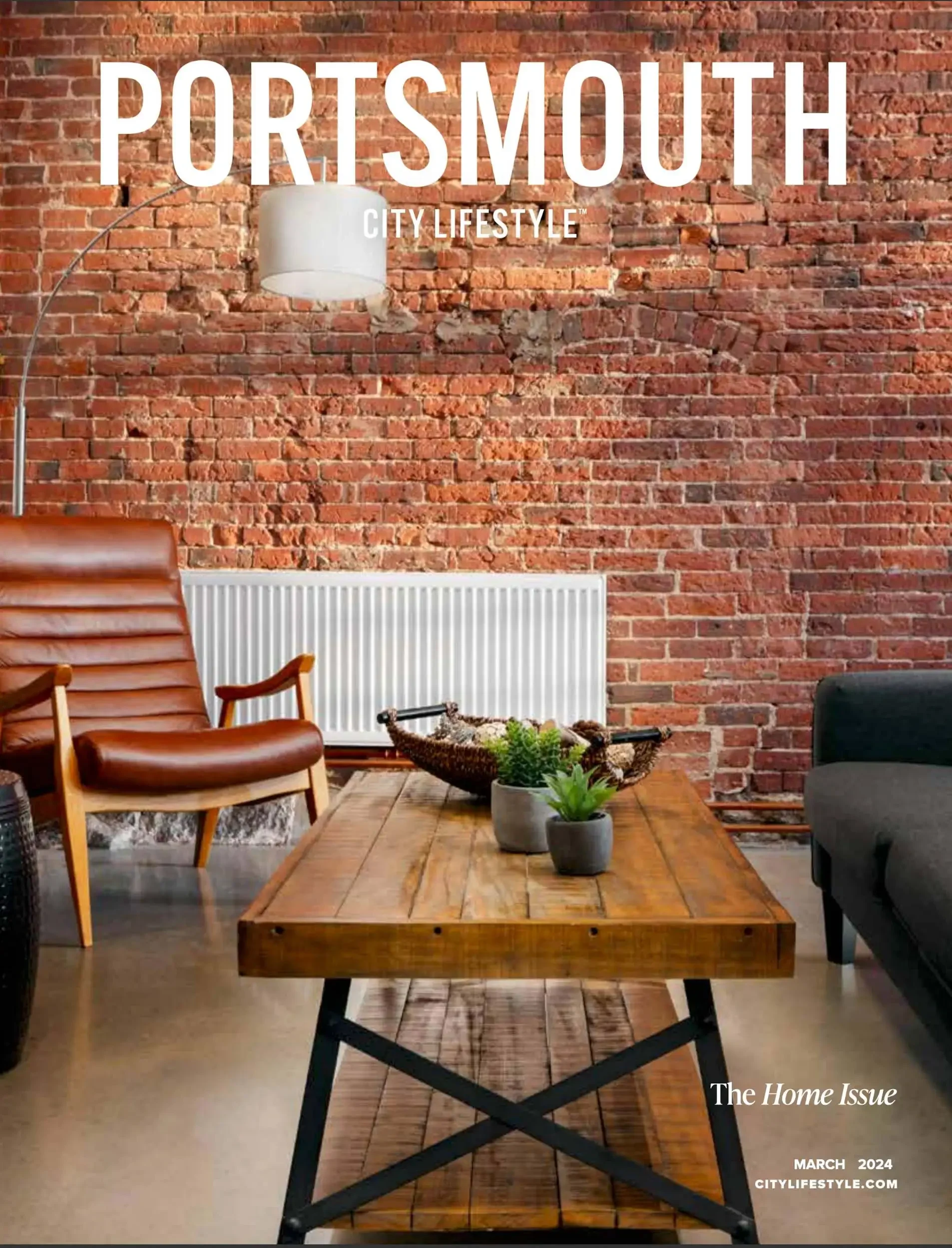 The cover of a magazine titled 'Portsmouth City Lifestyle' features a modern, cozy living space with a rustic brick wall, a wooden coffee table decorated with potted plants, a leather chair, a black sofa, a white radiator, and a white floor lamp, wit