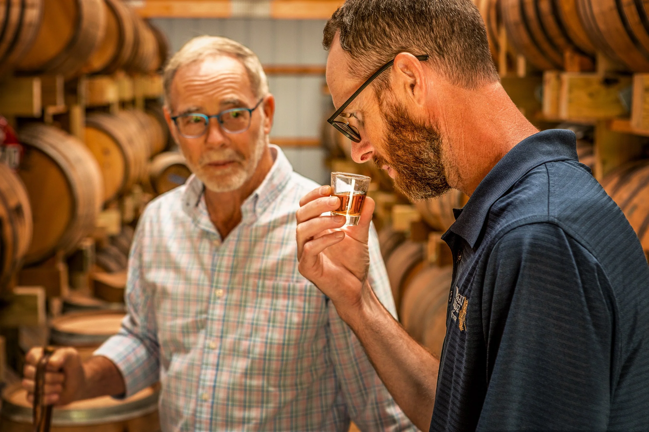 Two men in a spirits cellar. One man with a glass of whiskey, the other looking at him, surrounded by wooden barrels.