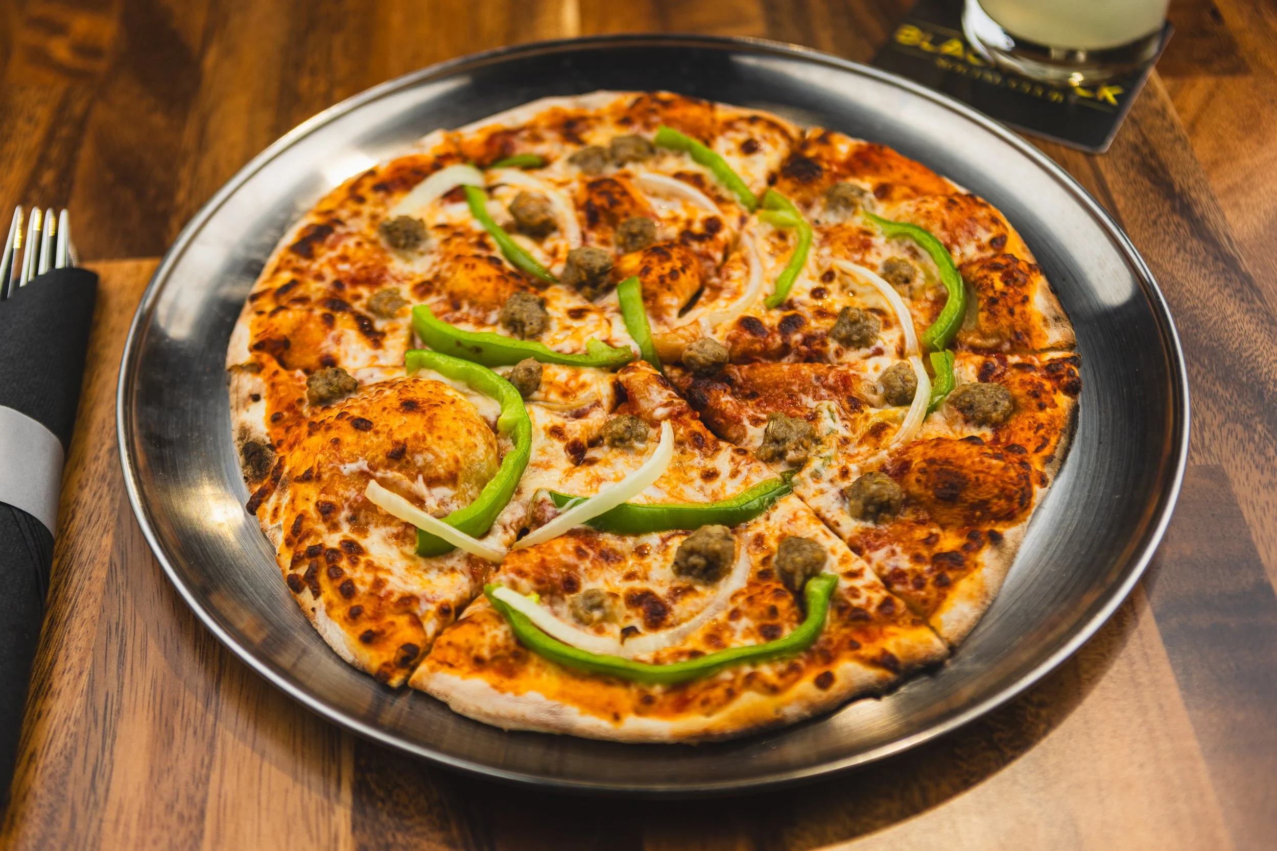 A pepperoni pizza topped with green bell peppers and sausage slices on a metal platter, placed on a wooden table.
