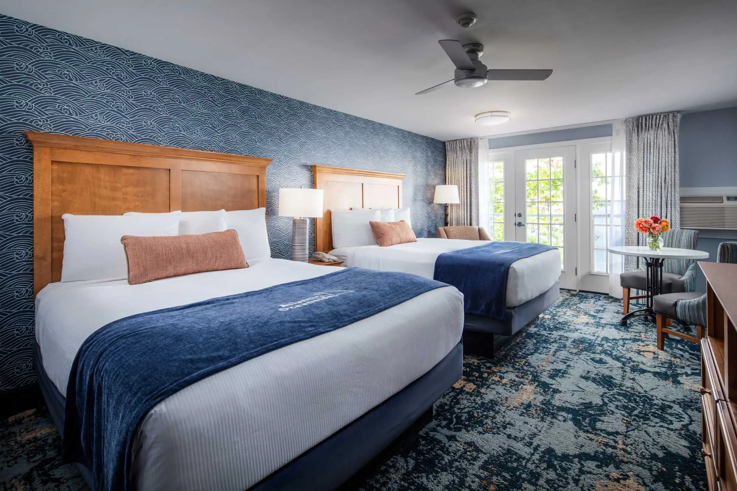 A hotel room with two beds, wooden headboards, white bedding, salmon-colored pillows, and dark blue throws. There are two bedside lamps and a window with sliding glass doors and curtains. A small table with a vase of pink and yellow flowers and a cha