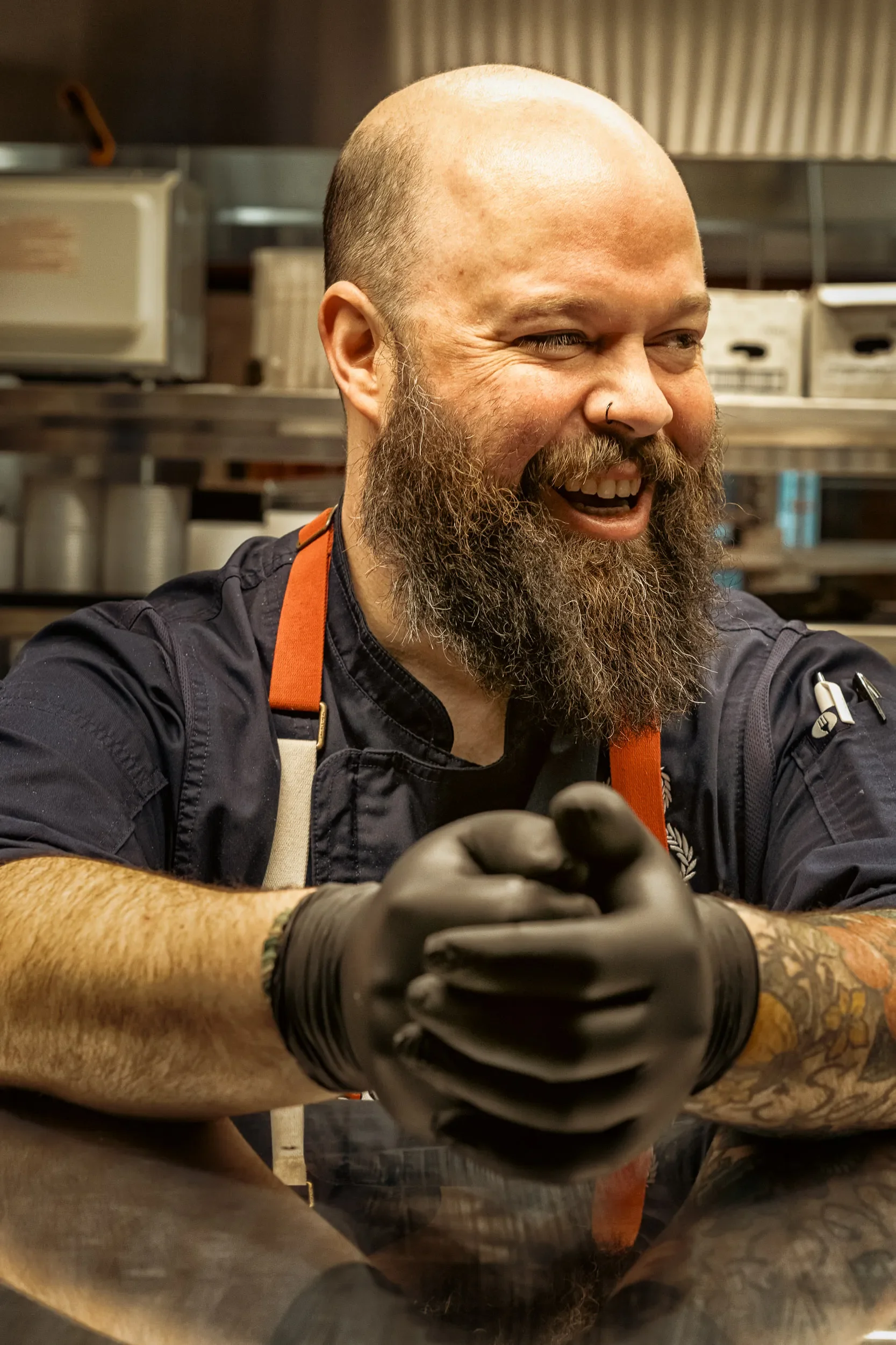 A smiling man with a beard, bald head, and a nose ring, wearing black gloves and a black shirt, working in a kitchen with stainless steel shelves and equipment in the background.