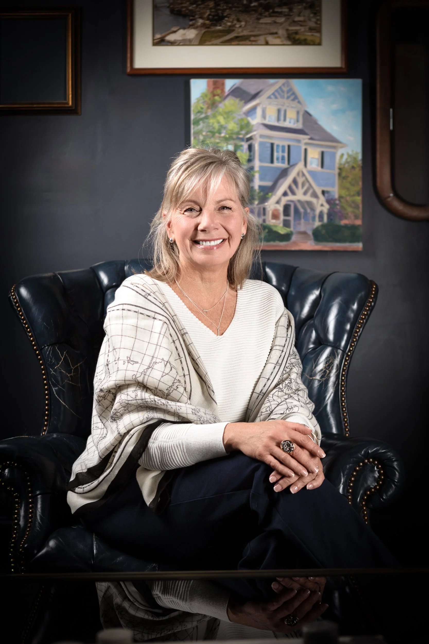 A smiling woman with blonde hair sits in a black leather armchair in a room with dark walls, with paintings of houses hanging behind her.