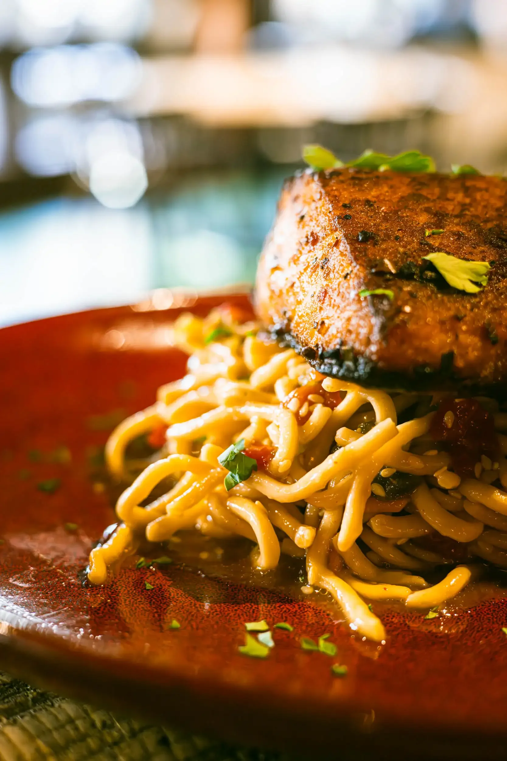 Close-up of a plate with noodles topped with a large piece of seasoned meat and garnished with chopped green herbs, in a warm indoor setting.