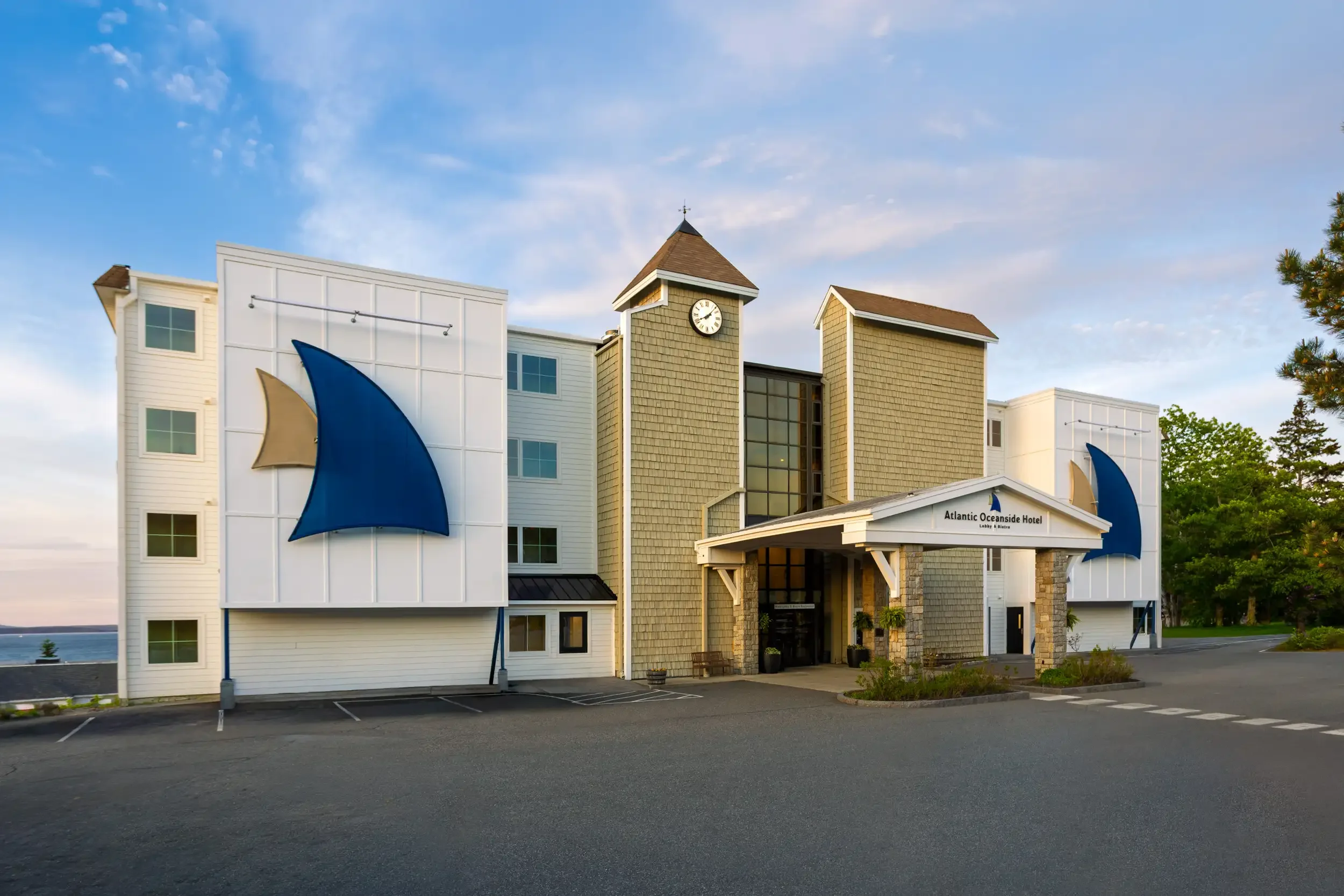 The Atlantic Oceanside Hotel features white, beige, and green exterior walls with a clock tower. Blue shark-shaped flags are on either side of the entrance. The sky is partly cloudy with a few trees visible on the right.