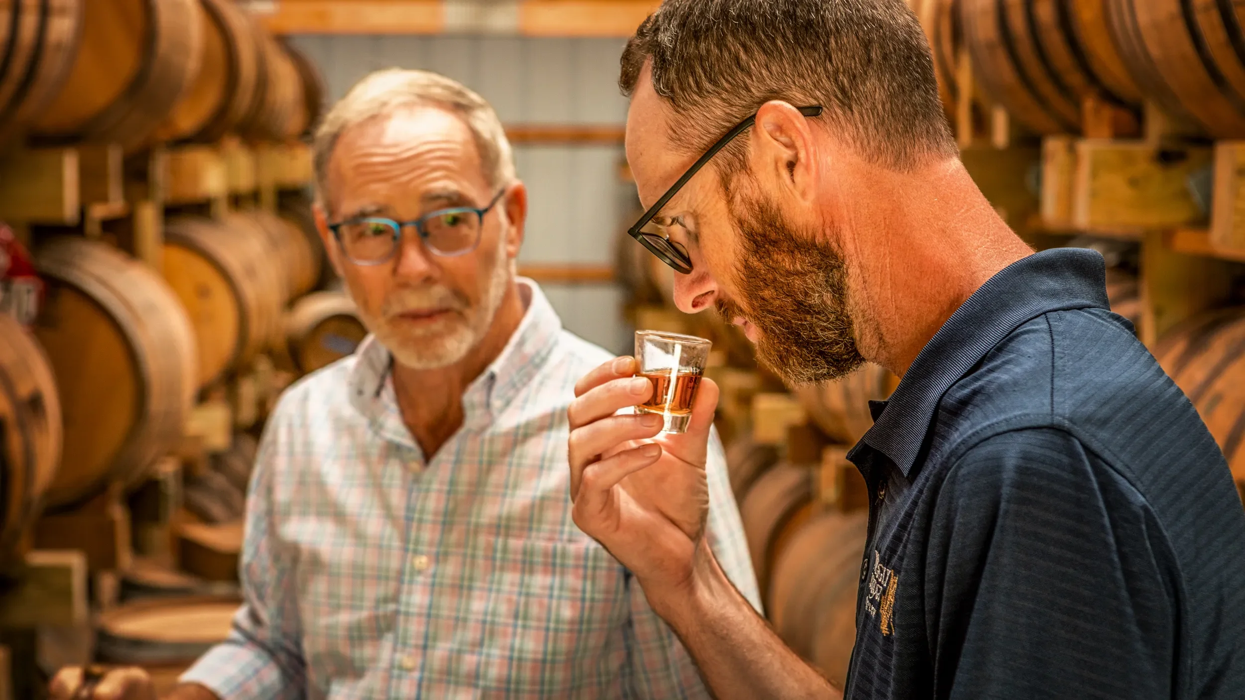 Two men in a bourbon warehouse, one older with gray hair and glasses, the other younger with a beard and glasses, holding a glass of bourbon, surrounded by wooden barrels.