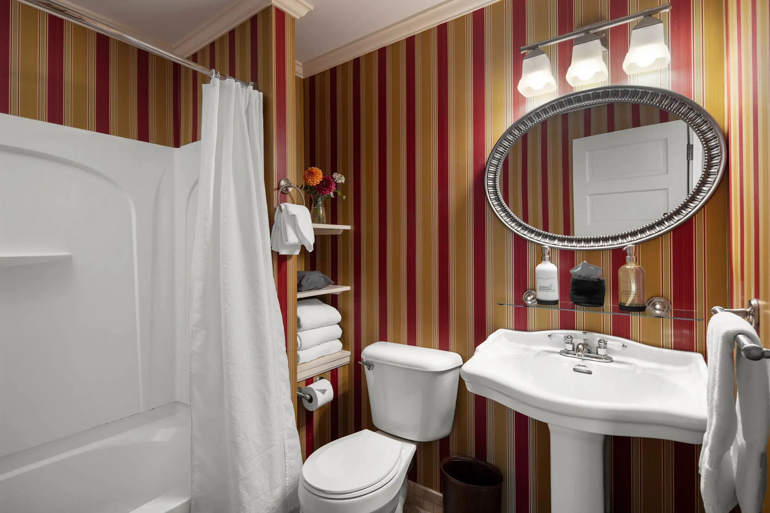 A small bathroom with striped red and yellow wallpaper, a white bathtub with white shower curtain, a toilet, a sink, and a mirror with three lights above.