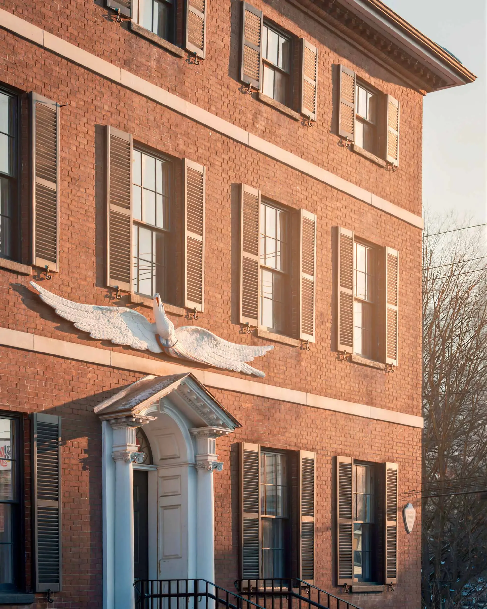 The exterior of a red brick building with multiple windows featuring brown shutters, and a decorative white swan sculpture mounted on the wall below the windows. There is a small entryway with a white decorative arch and railing, and some bare tree branches are visible on the right side of the image.