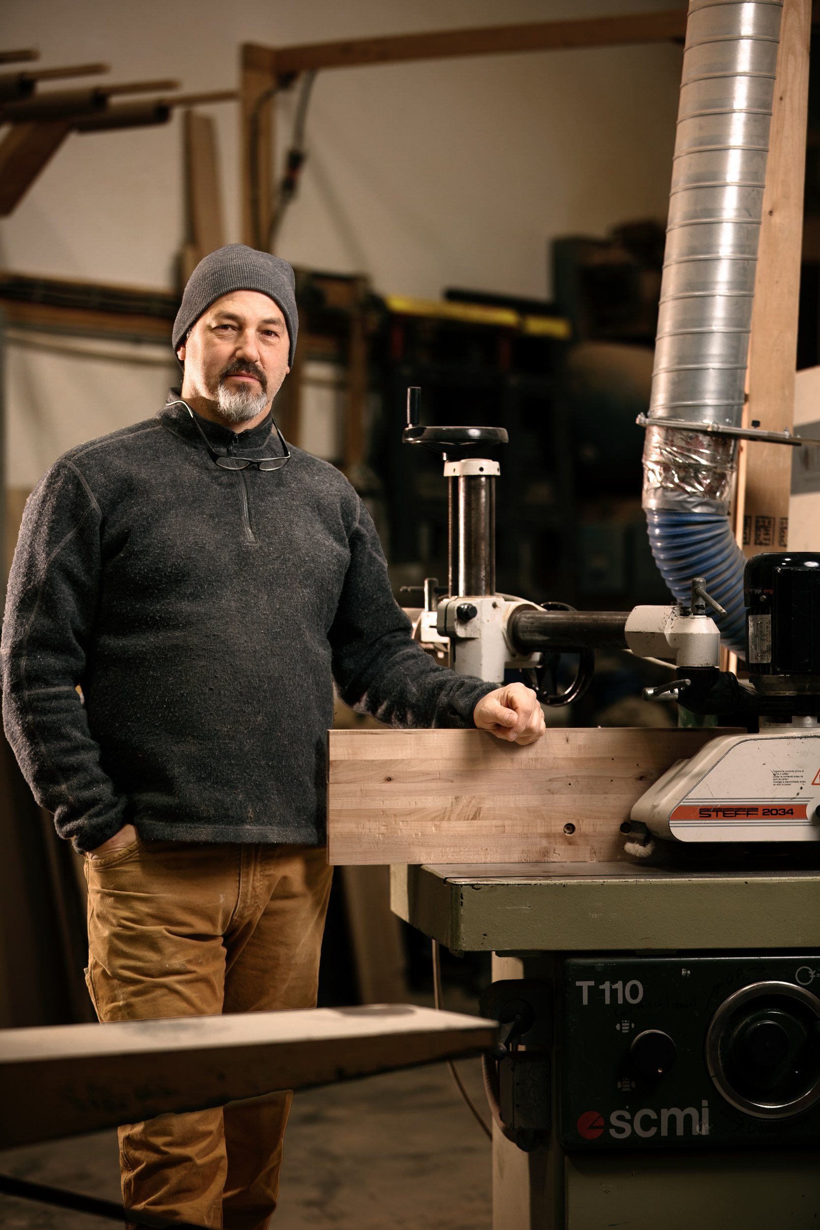Man in a dark gray sweater and beanie standing next to a woodworking machine in a workshop.